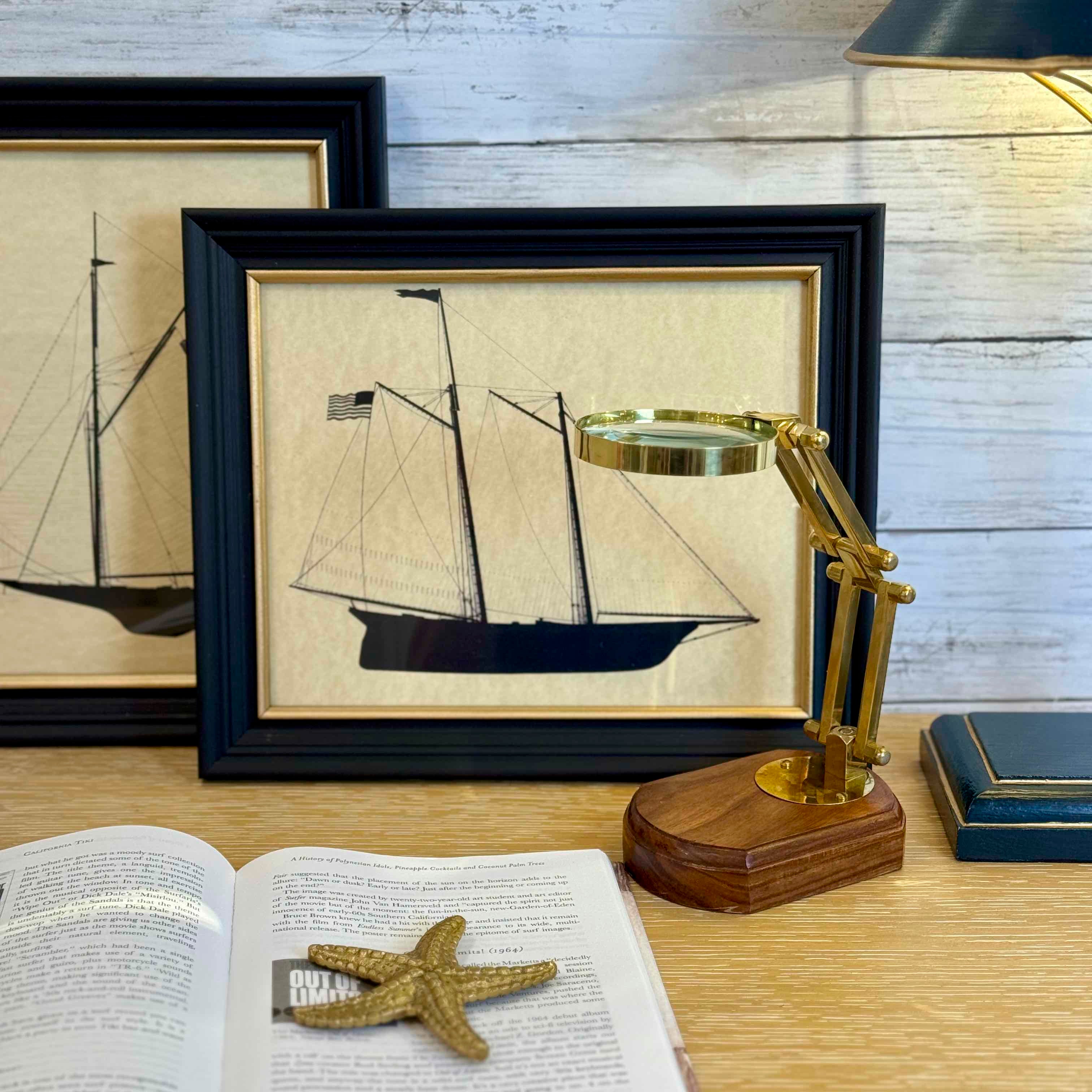 Framed illustration of a ship on a wooden surface with a magnifying glass and open book.