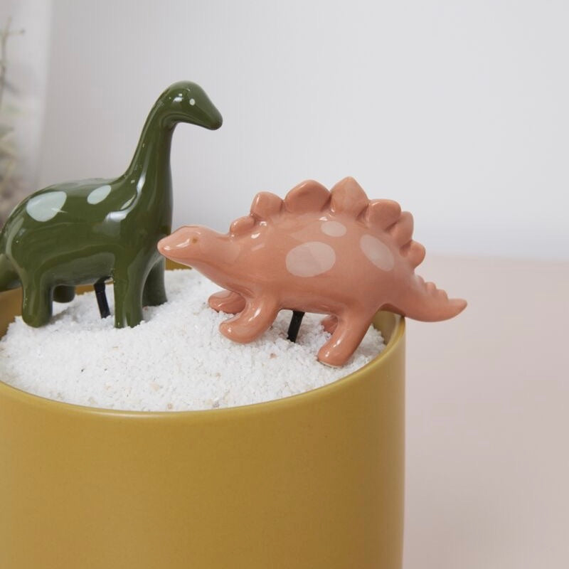 Two ceramic dinosaur figures, one green and one pink, in a container of sand on a white background.