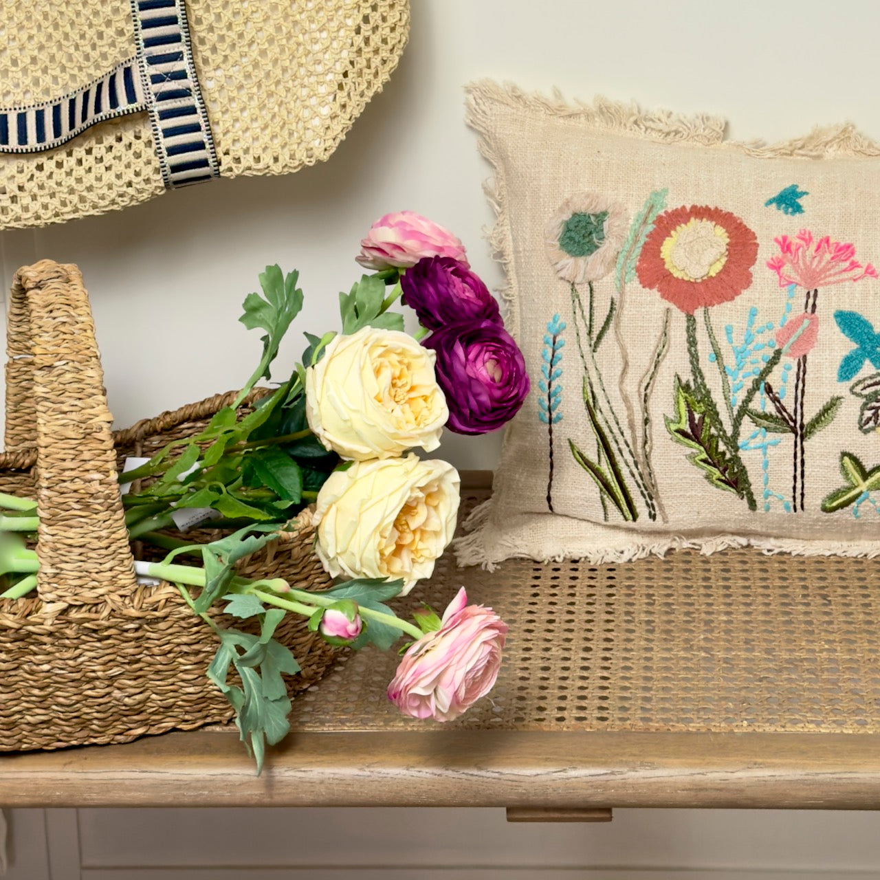 Decorative setting with a straw hat, basket of flowers, and embroidered pillow on a wooden surface.