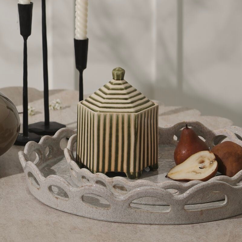 A ribbed ceramic lidded jar in beige and olive green sits on a scalloped light gray tray with pear slices and black candlesticks in the background.
