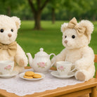 Two teddy bears with bows sitting at a tea set on a table outdoors.