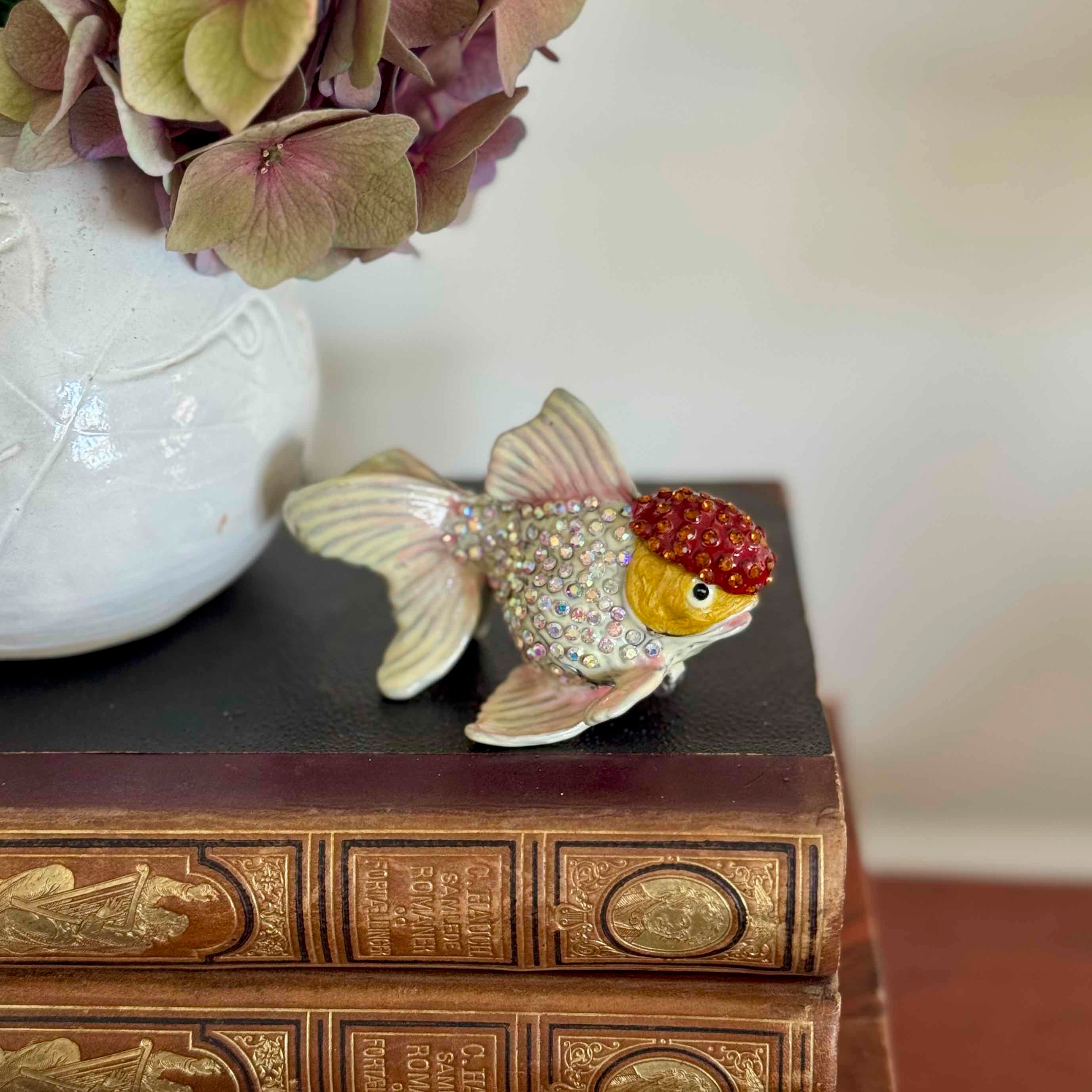 Decorative fish figurine with a red gem on a book next to a white vase with flowers.