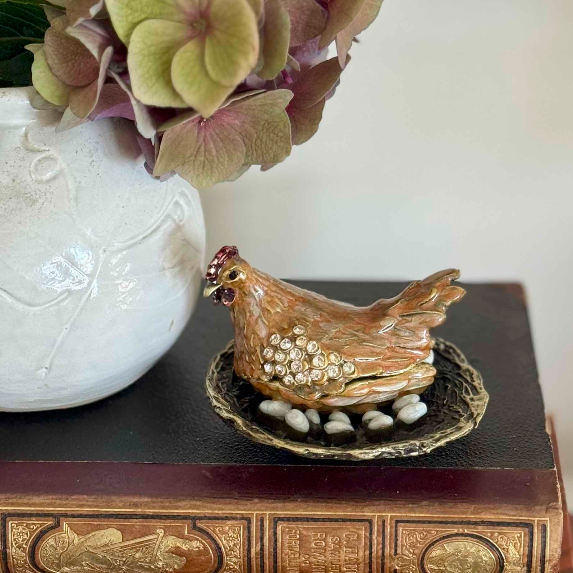 Decorative ceramic chicken with eggs on a stand next to a white vase with flowers.