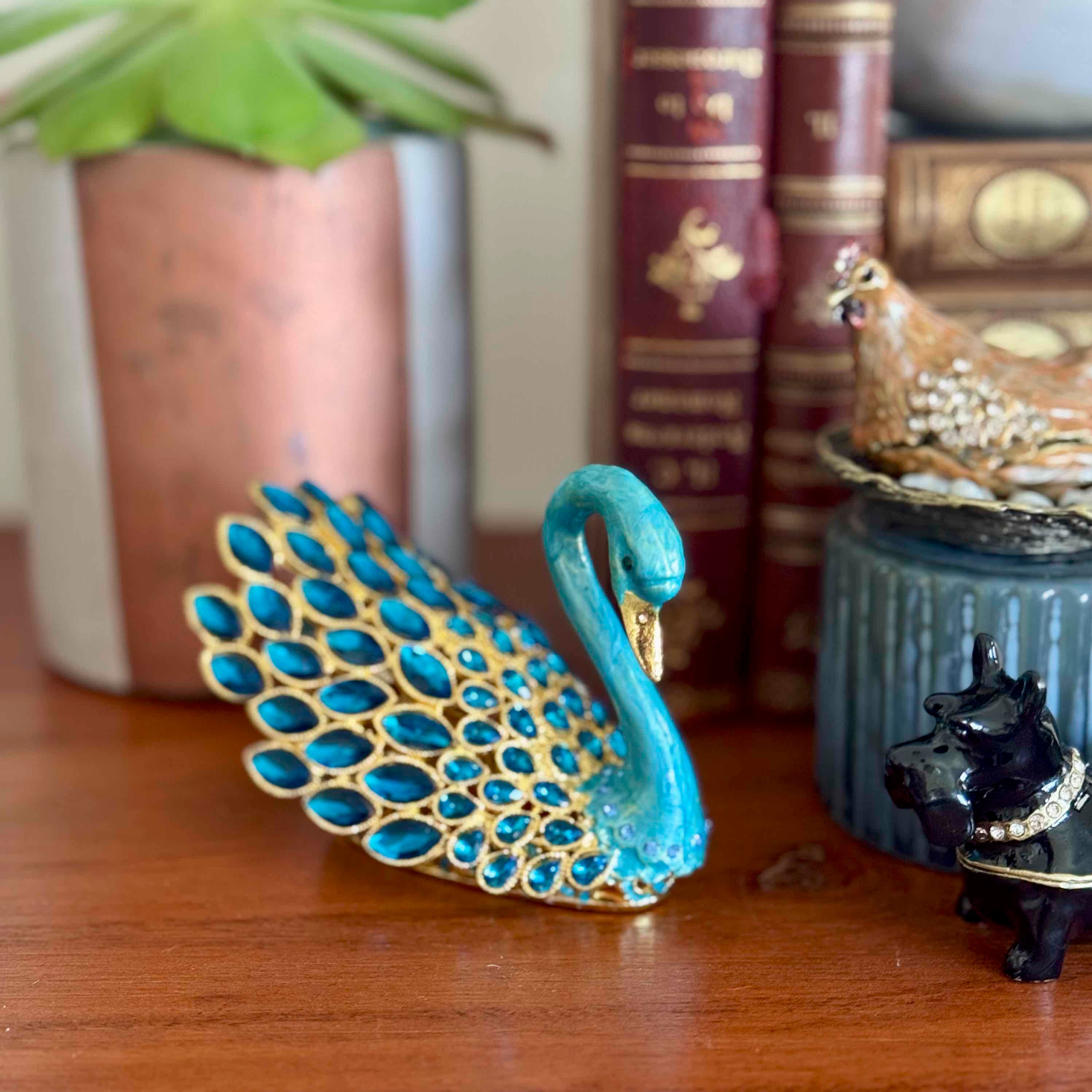 Decorative peacock figurine on a wooden surface with books and other small objects in the background.