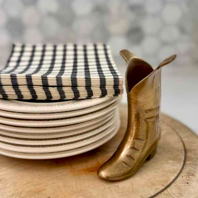 Brass boot-shaped container next to stacked plates with a striped cover on a wooden surface.