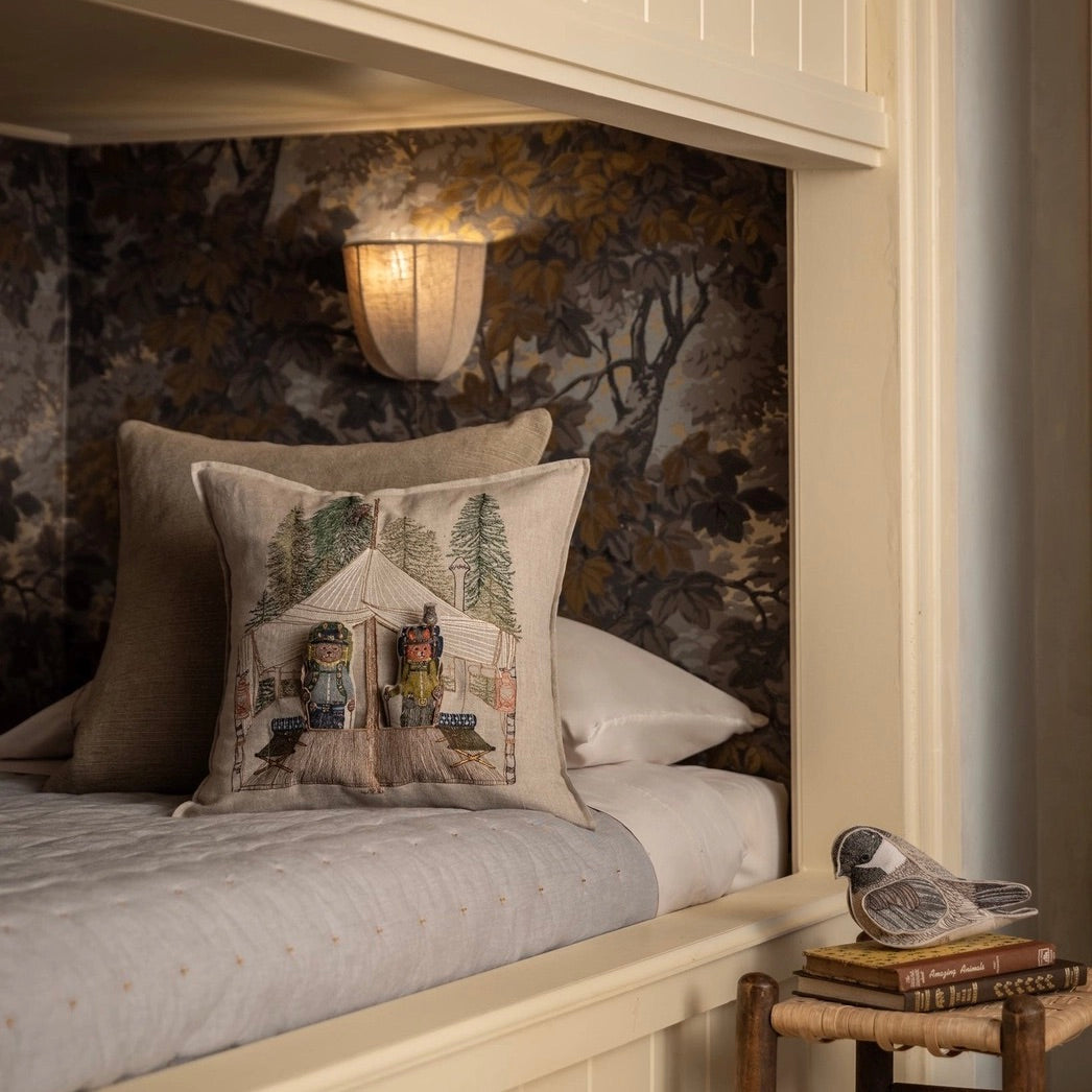 A styled bedroom corner featuring a bed with a decorative throw pillow printed with a vintage camping scene. The background is decorated with dark, moody botanical wallpaper and a warm wall sconce.