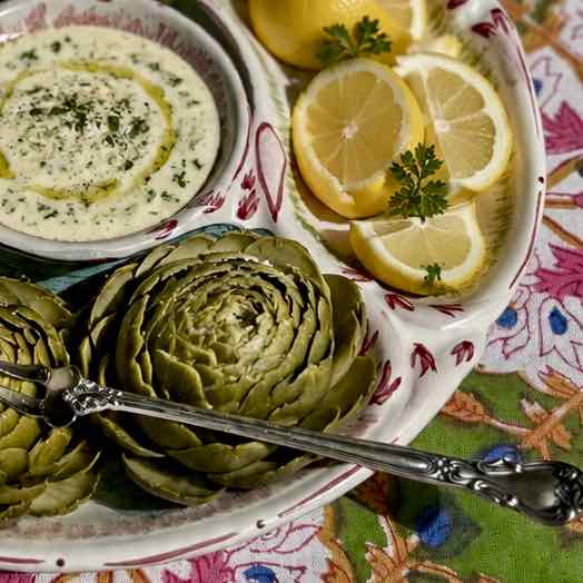 A close-up of a serving platter holding two green artichokes, a small bowl of white dip with herbs and olive oil, and lemon slices on a patterned tablecloth.