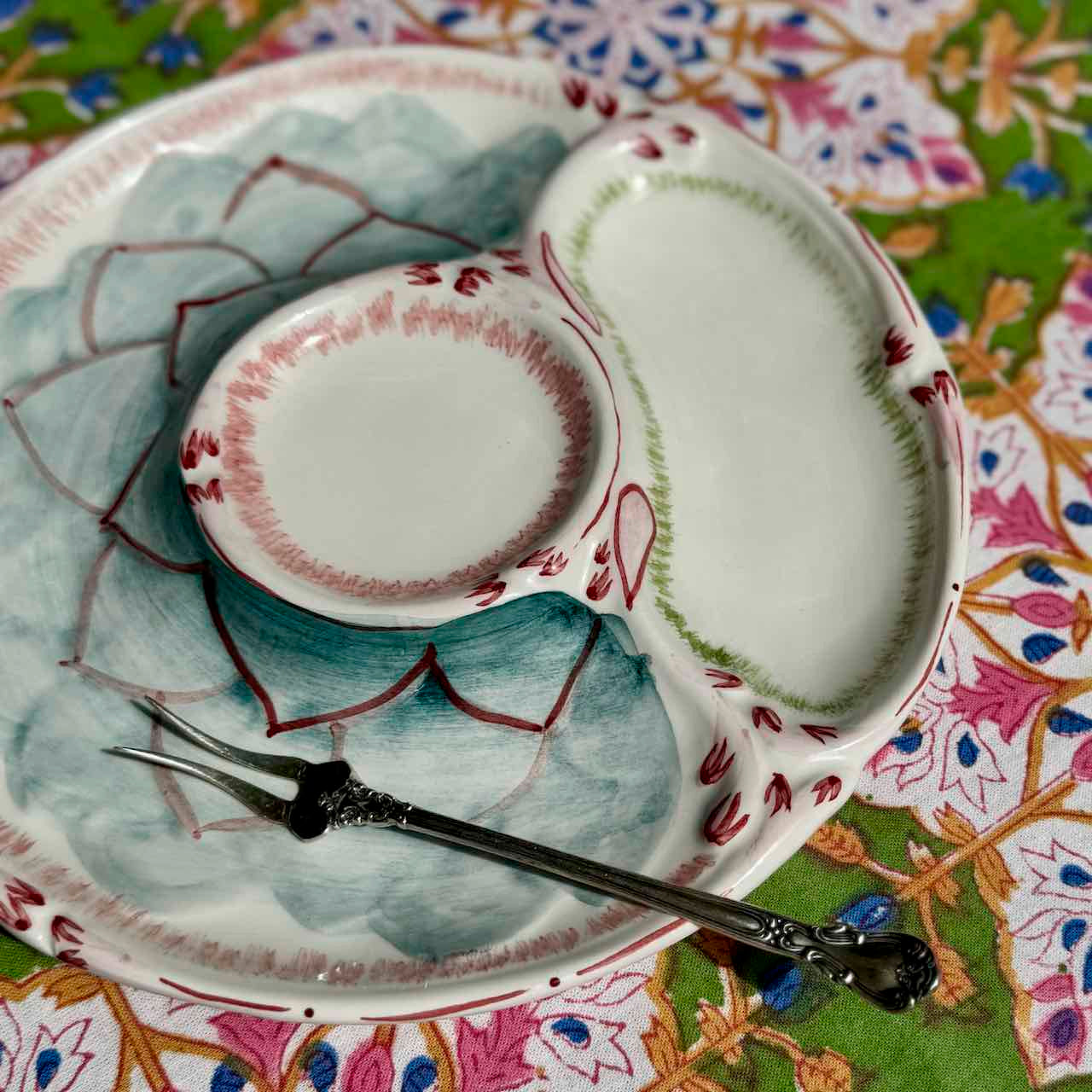 A ceramic serving platter with three compartments, decorated with a hand-painted lotus motif in shades of green, blue, and red, accompanied by a silver serving fork, set on a colorful patterned textile.