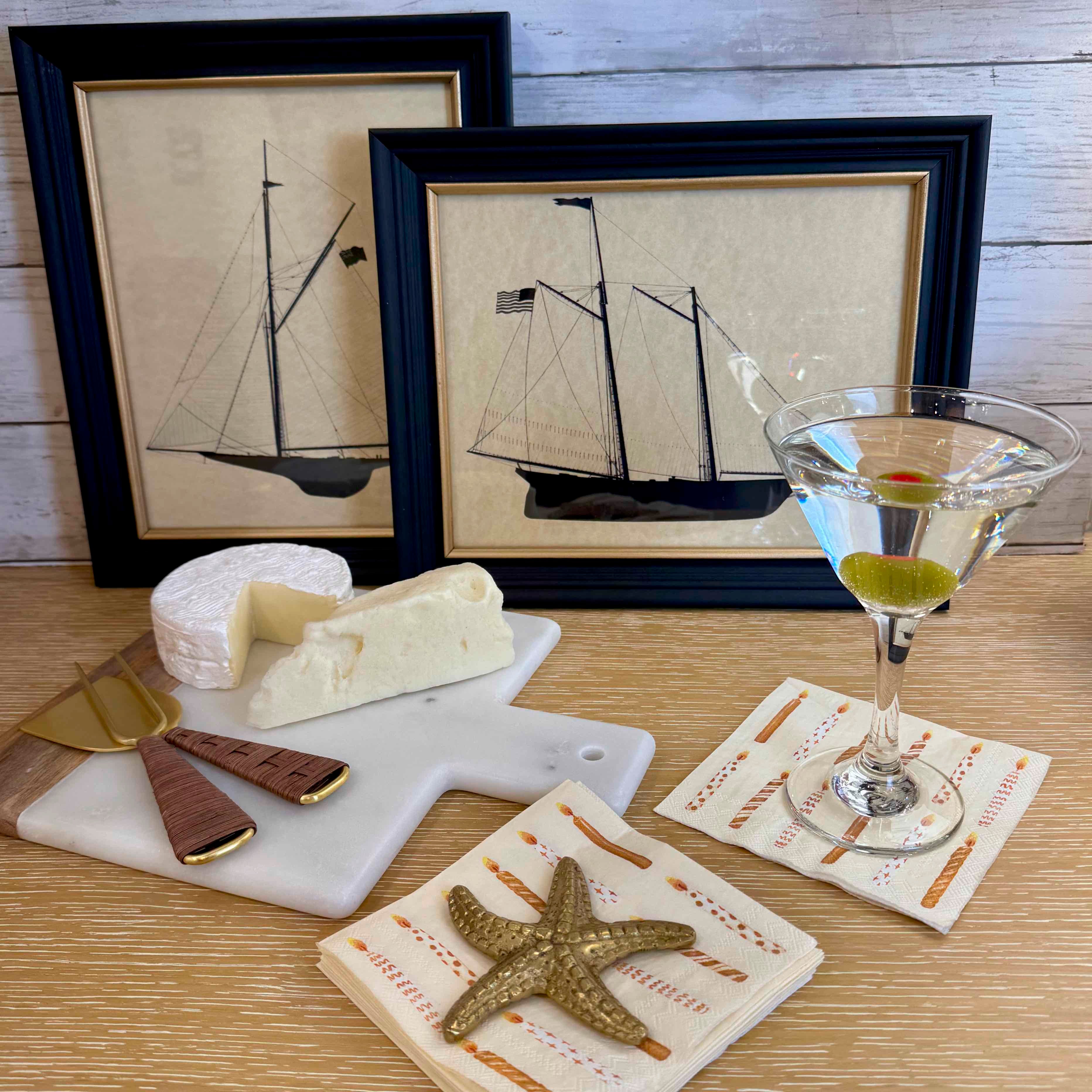 Table setting with cheese, chocolate, a starfish, and a martini glass in front of framed ship artwork.
