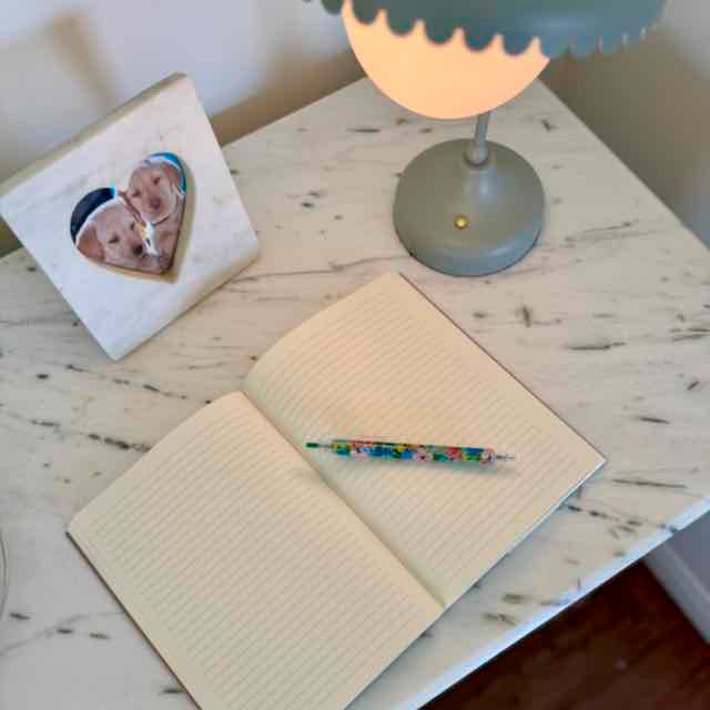 Open notebook with a pen on a wooden desk next to a lamp and photo frame.