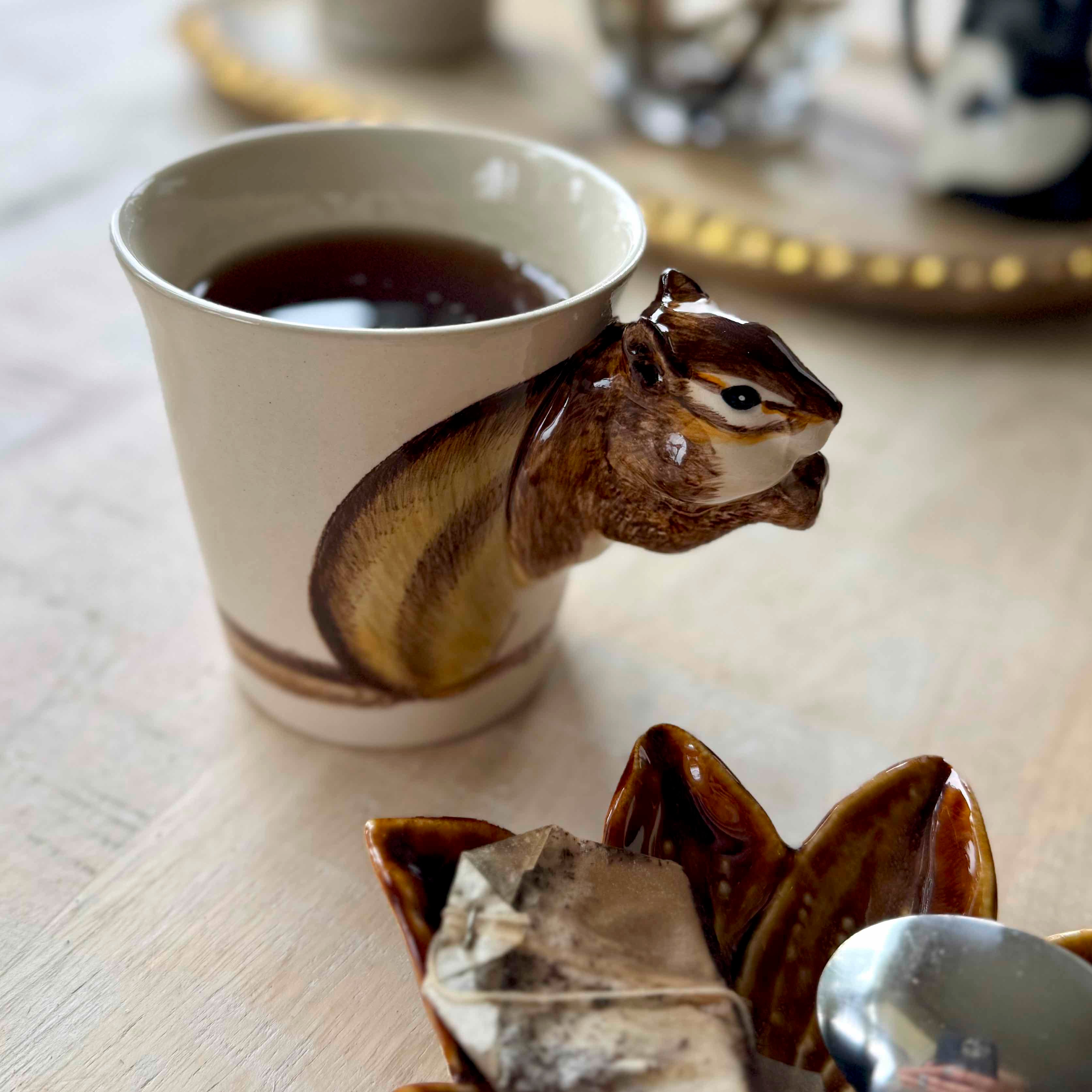 Squirrel-shaped mug with a drink on a wooden surface with tea bags and a spoon.