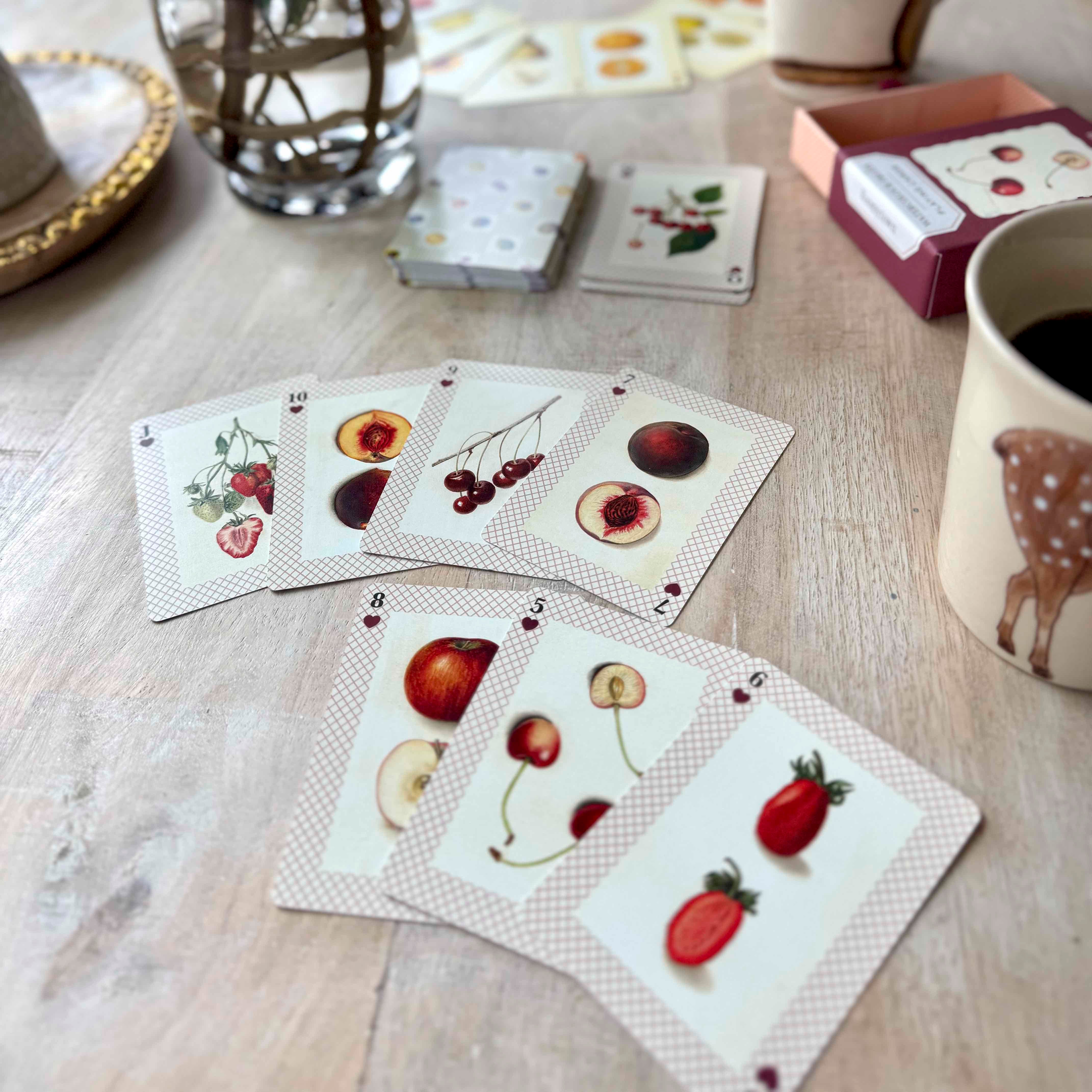 Playing cards with fruit illustrations on a wooden table with a mug and glass.