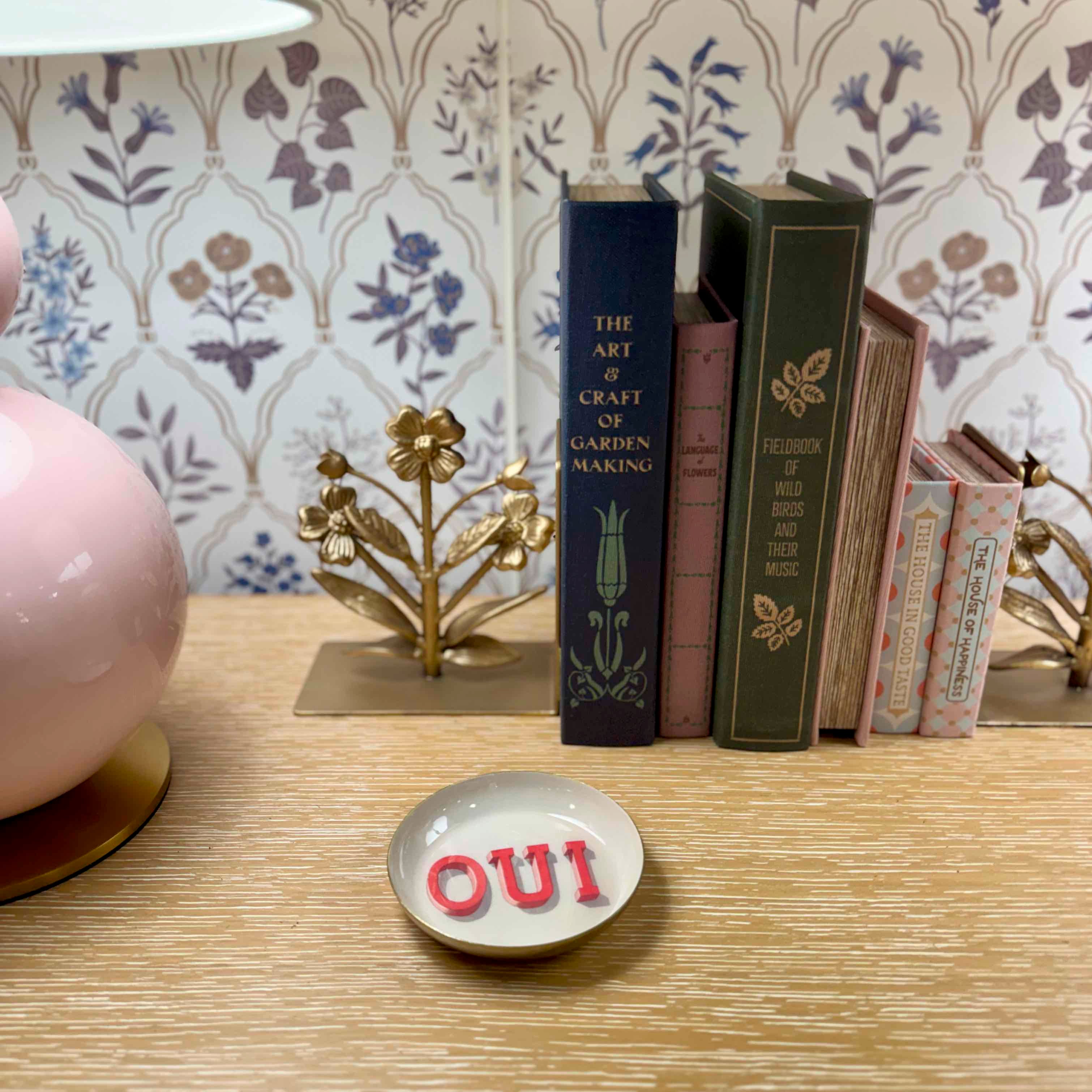 Tabletop scene with books, a decorative item, and a small dish with 'OUI' letters on a patterned wall background.
