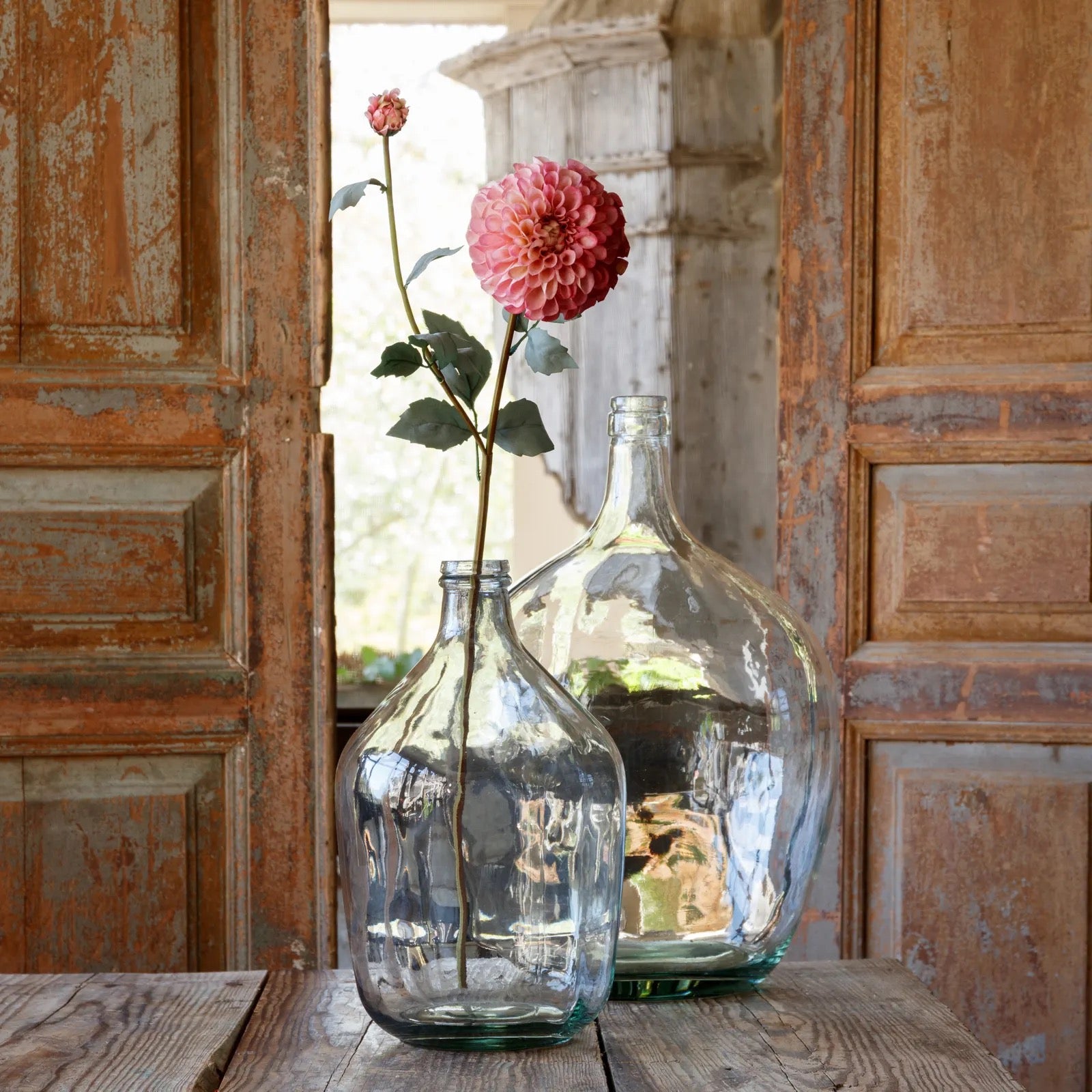 Two glass bottles with a single pink flower against a rustic wooden door background