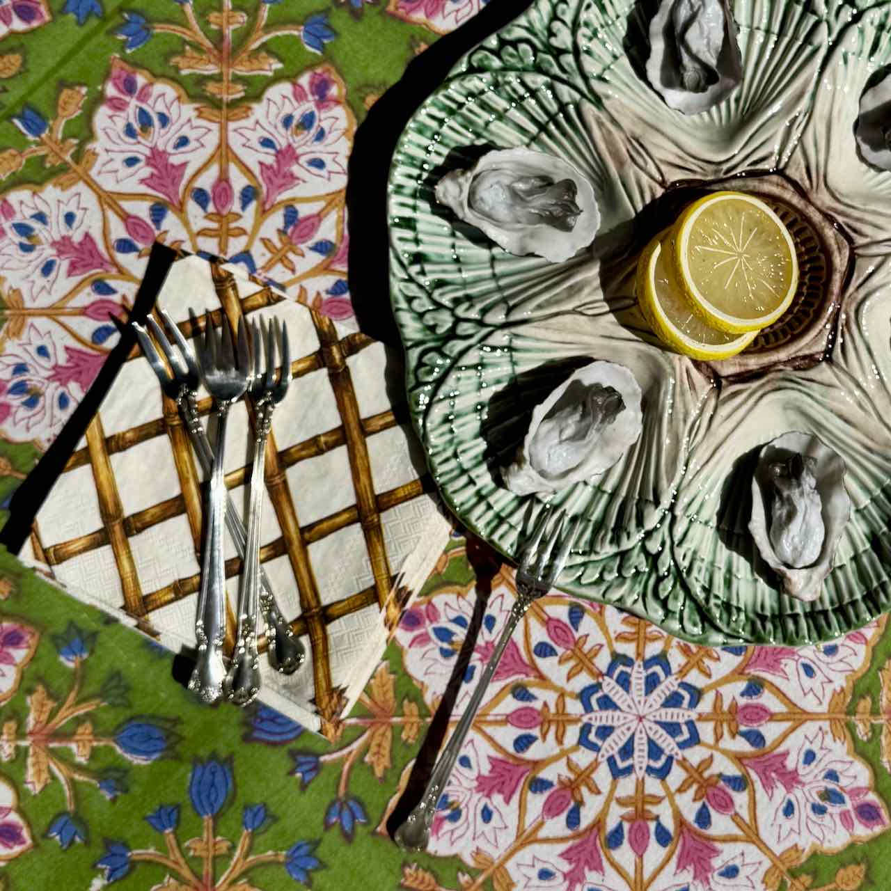 An overhead styled shot of oysters served on a green scalloped ceramic platter with lemon slices, alongside silver forks on a patterned tablecloth.