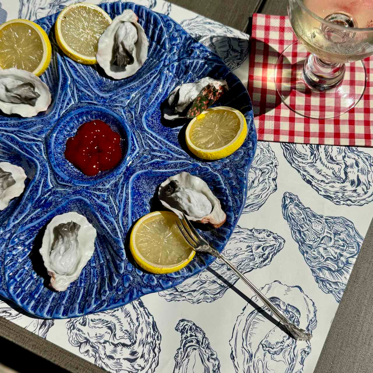 A blue ceramic oyster platter with lemon slices and a small red dipping sauce, set on a patterned tablecloth next to a red-and-white checkered napkin and a wine glass.