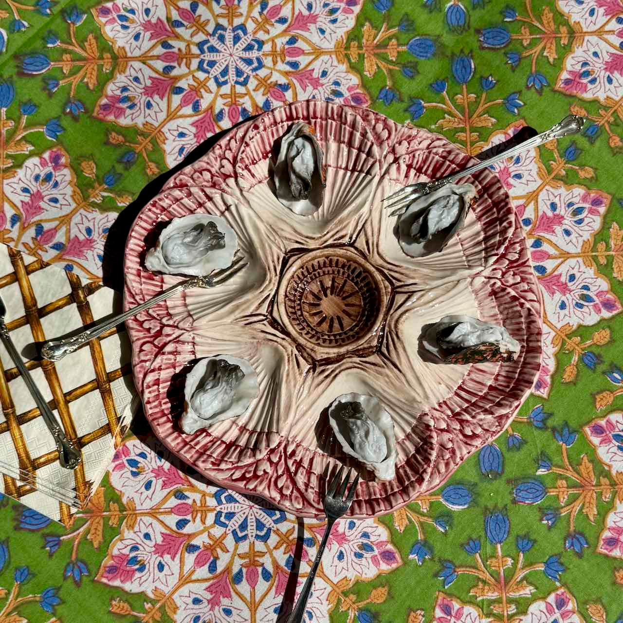 A ceramic oyster shell serving platter with six carved oyster shells, set on a vibrant green and pink patterned tablecloth with forks and a small wooden trivet.