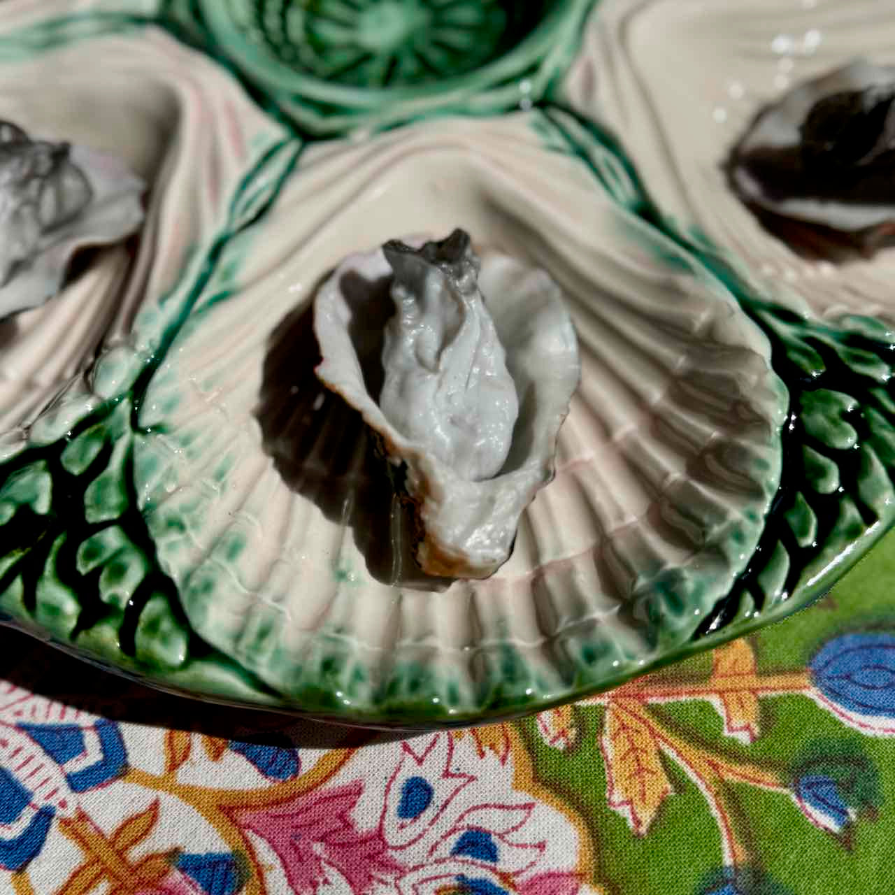 A close-up of a ceramic oyster shell serving dish holding several raw oysters, set on a colorful floral-patterned fabric surface.