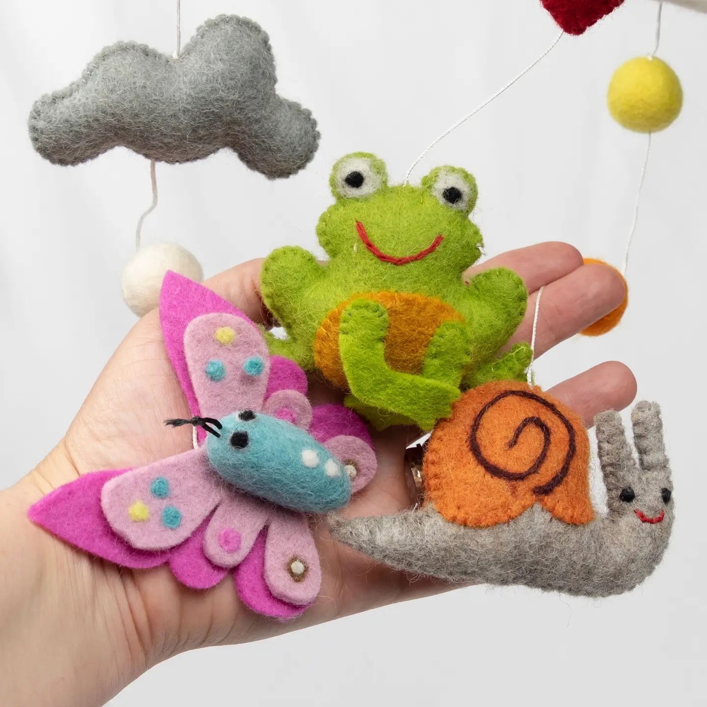 Hand holding felt animal toys including a frog, butterfly, and snail against a white background.