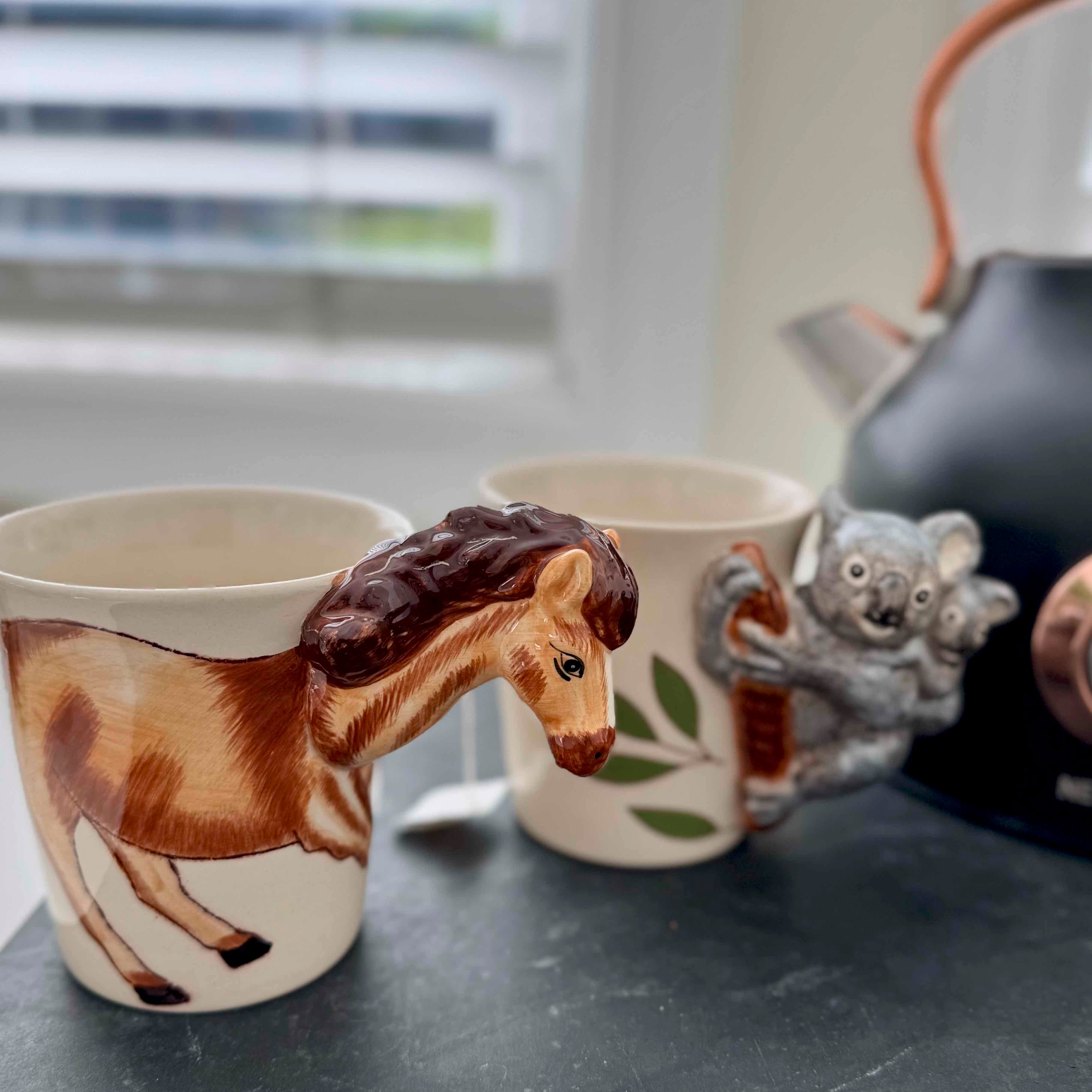 Mug with horse design, another mug with koala design, and a teapot on a surface.