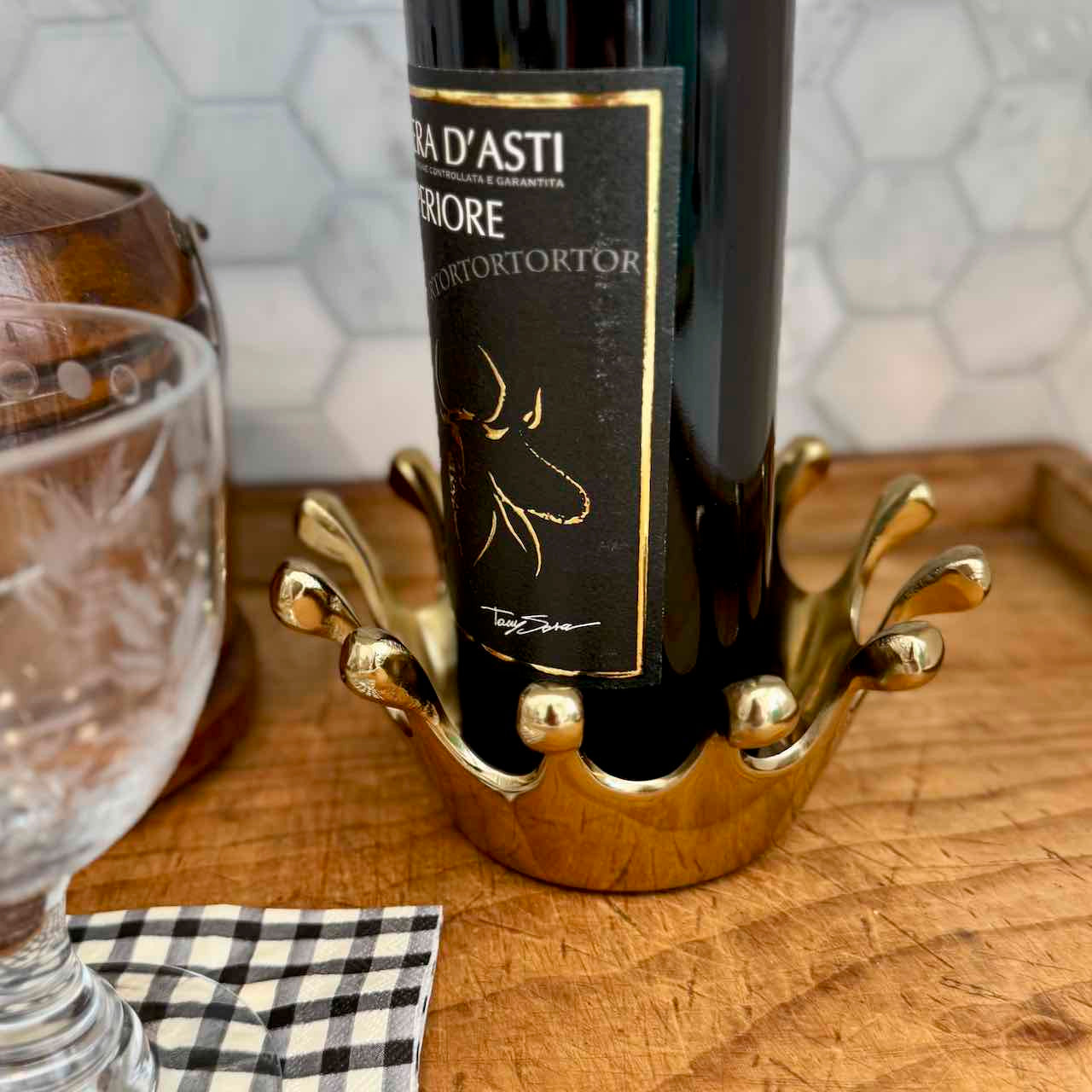 A gold crown-shaped wine bottle holder cradling a dark wine bottle on a wooden tray, with a glass and a checkered napkin nearby.
