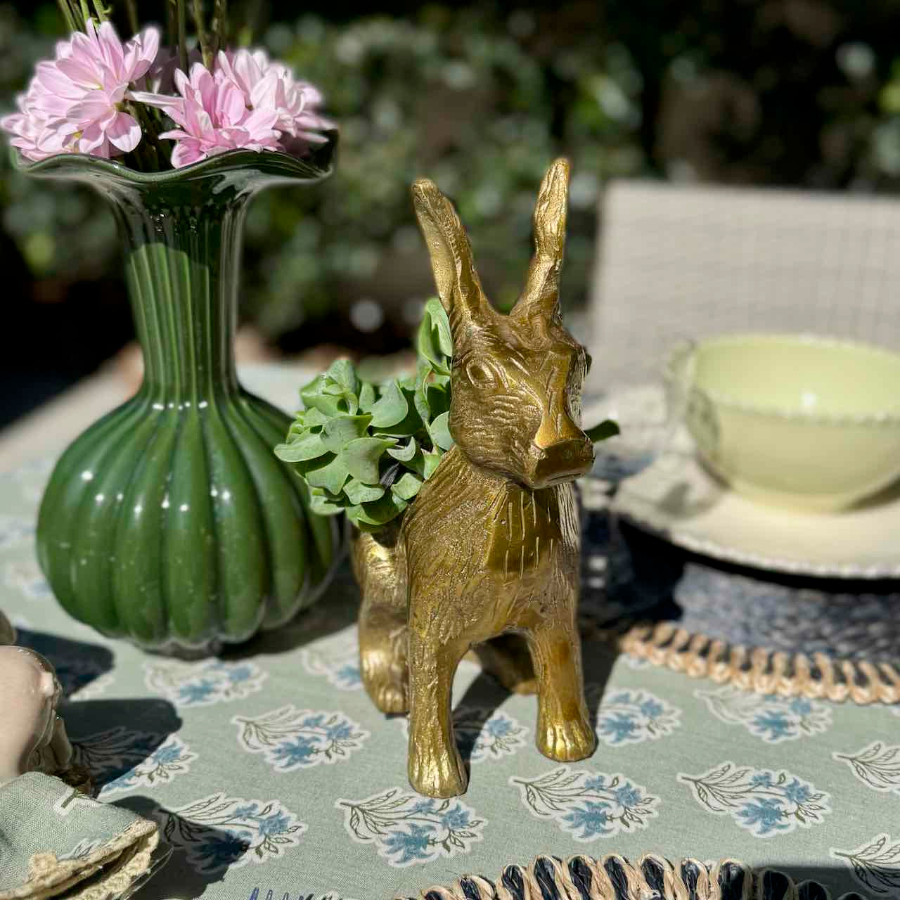 Gold rabbit-shaped vase on a table with a green vase and flowers in the background