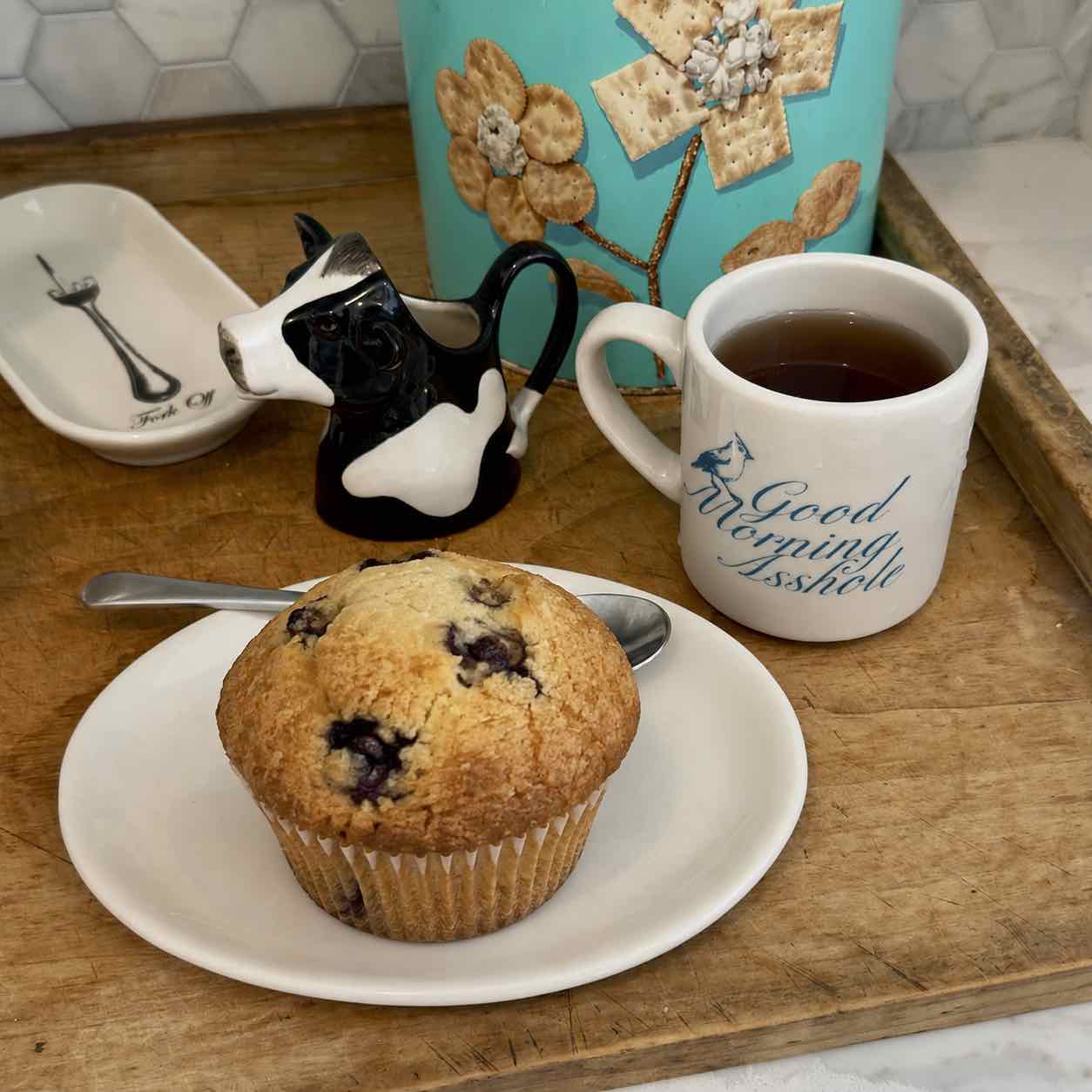 A breakfast scene on a wooden tray with a blueberry muffin on a white plate, a white mug with blue script text, a small black-and-white cow-shaped pitcher, and a turquoise decorative container in the background.