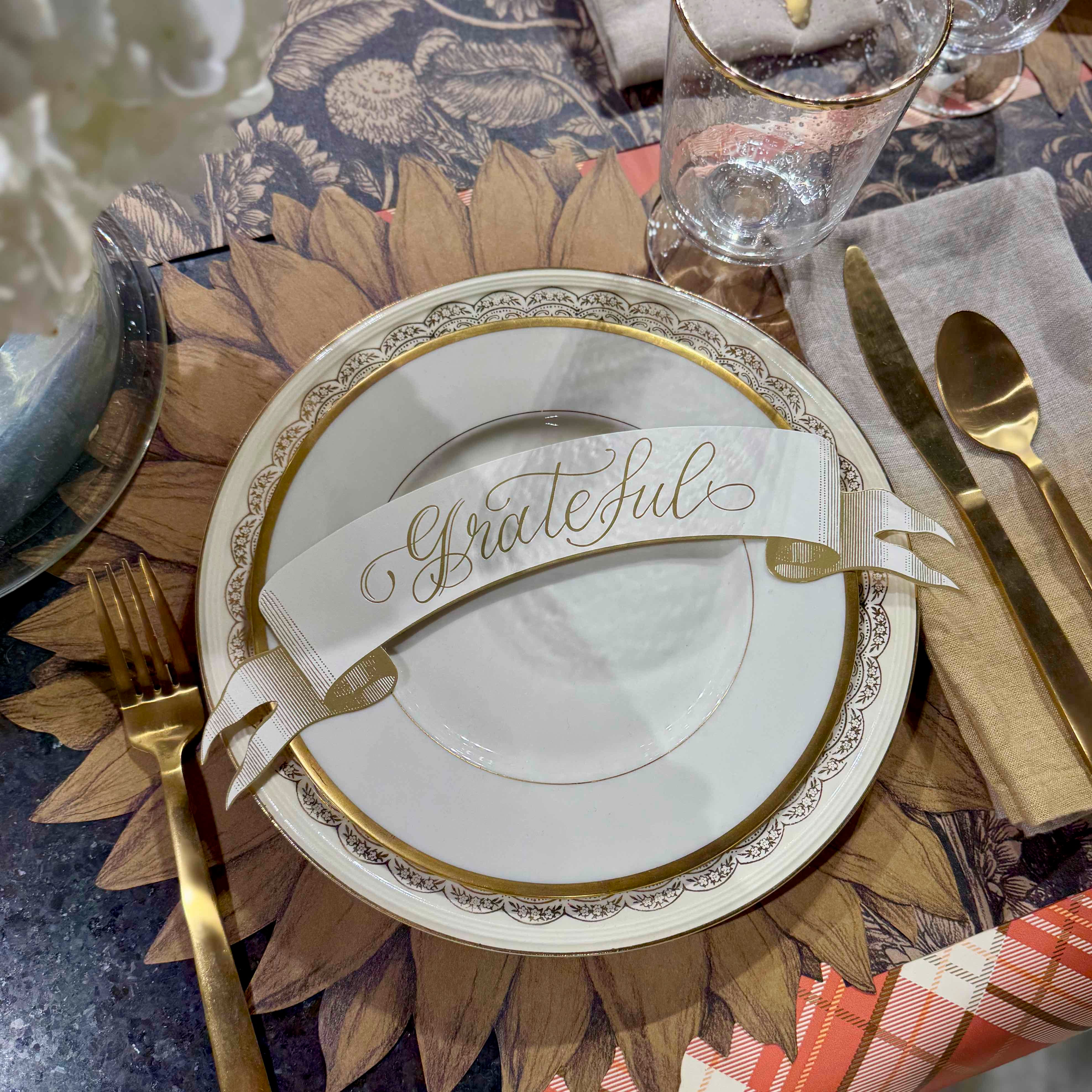 Decorative table setting with a plate featuring the word 'Grateful', gold cutlery, and a patterned tablecloth.