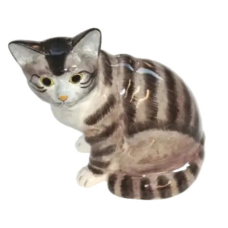 A ceramic cat figurine with gray, black, and white tabby stripes, featuring yellow eyes and a small orange nose, shown on a plain light background.
