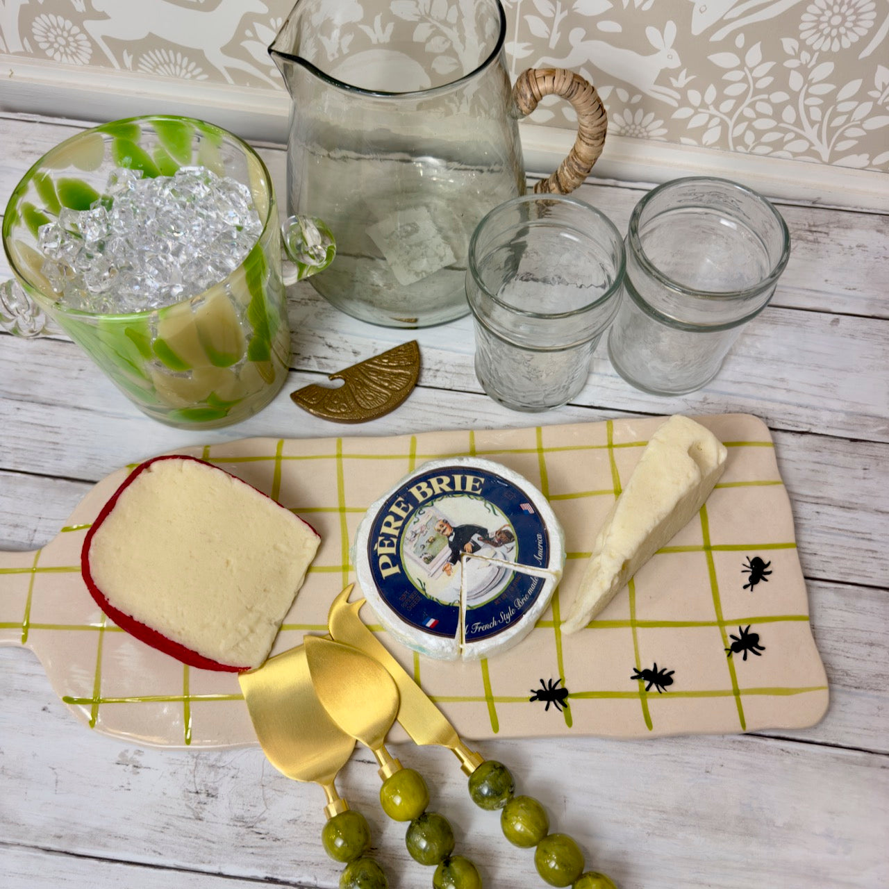 Cheese platter with cheeses, a jar of pickles, and glasses on a wooden surface.
