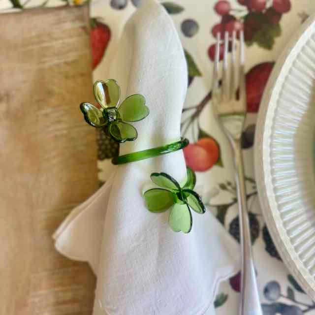 Green clover-shaped napkin ring on a white napkin with a floral-patterned plate in the background.