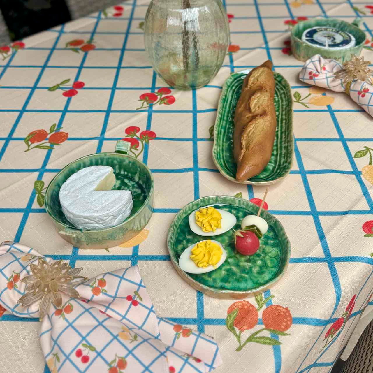 A styled tabletop scene with a cream tablecloth printed with a blue grid and red fruit motifs. On the table are green ceramic plates and bowls holding a sliced white cheese, a baguette, hard-boiled eggs with yellow yolks, a small red cherry, and folded patterned napkins.