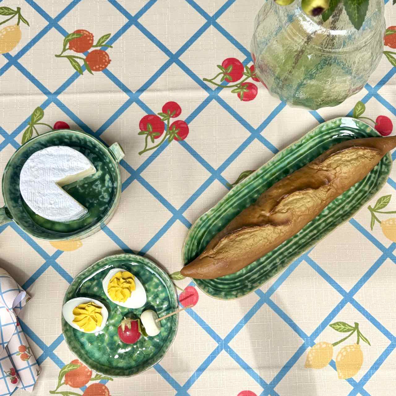 A styled overhead flat-lay of ceramic tableware on a printed tablecloth. The set includes a green speckled mug with a slice of white cake, a green oval platter holding a loaf of bread, a green round plate with halved hard-boiled eggs and small red and yellow accents, and a small green dish with a striped handle.