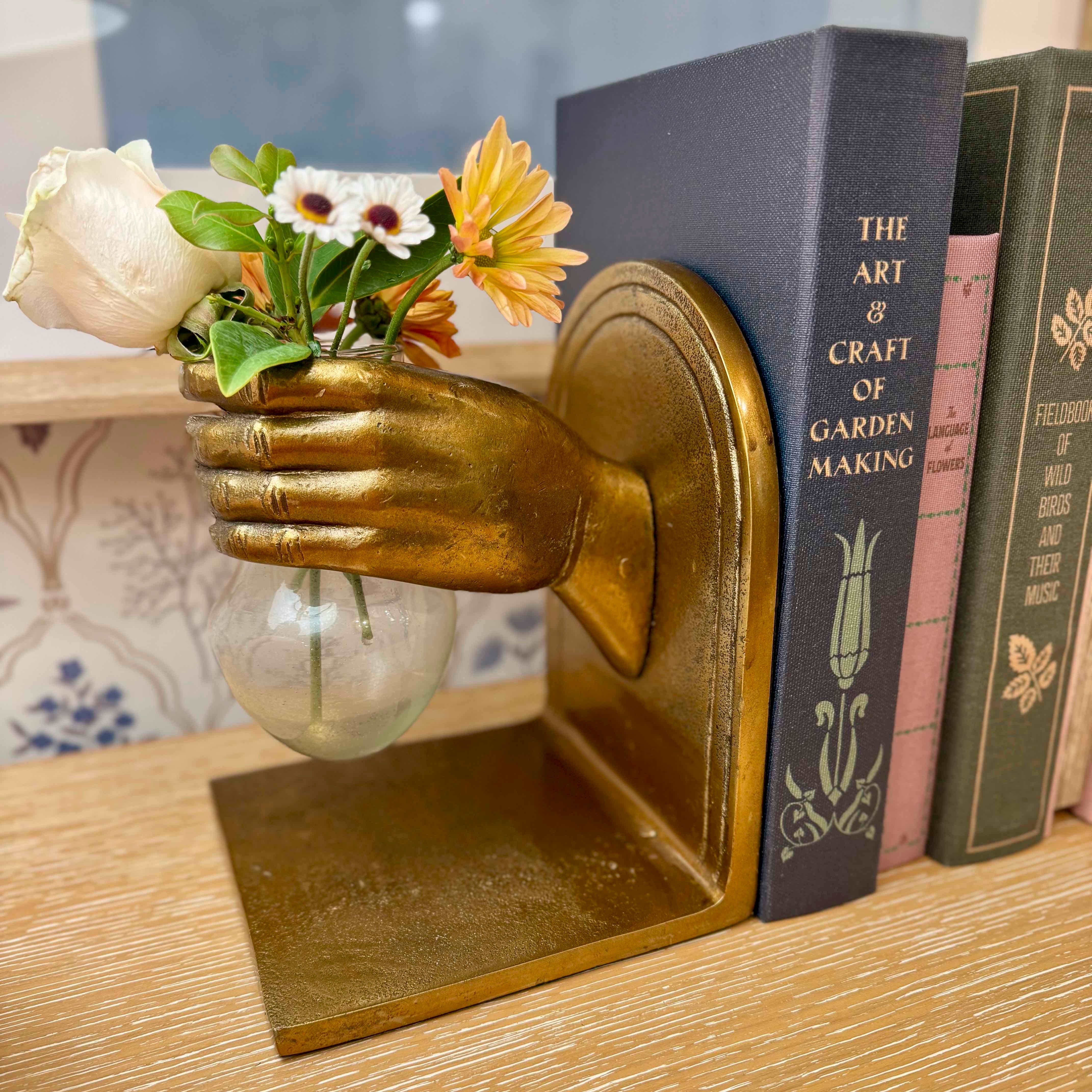Gold hand-shaped bookend with a small vase holding flowers, placed on a wooden surface with books in the background.