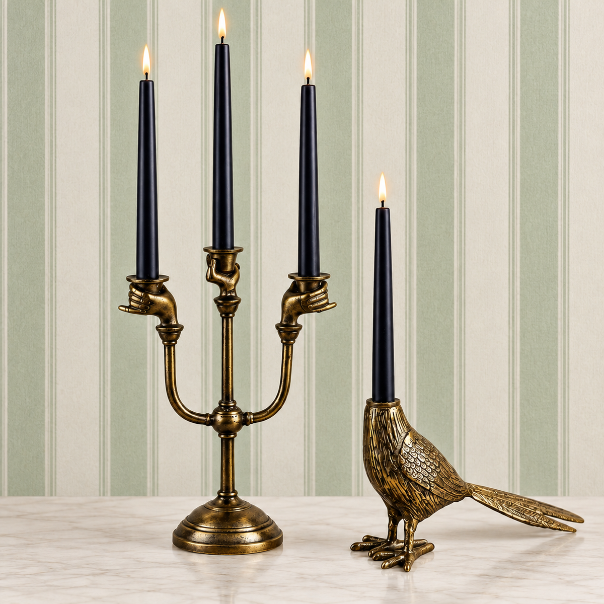 Two ornate brass candle holders on a light marble surface against a striped wallpaper background. One holder features three curved arms held by hands and holds three black taper candles lit at the top.