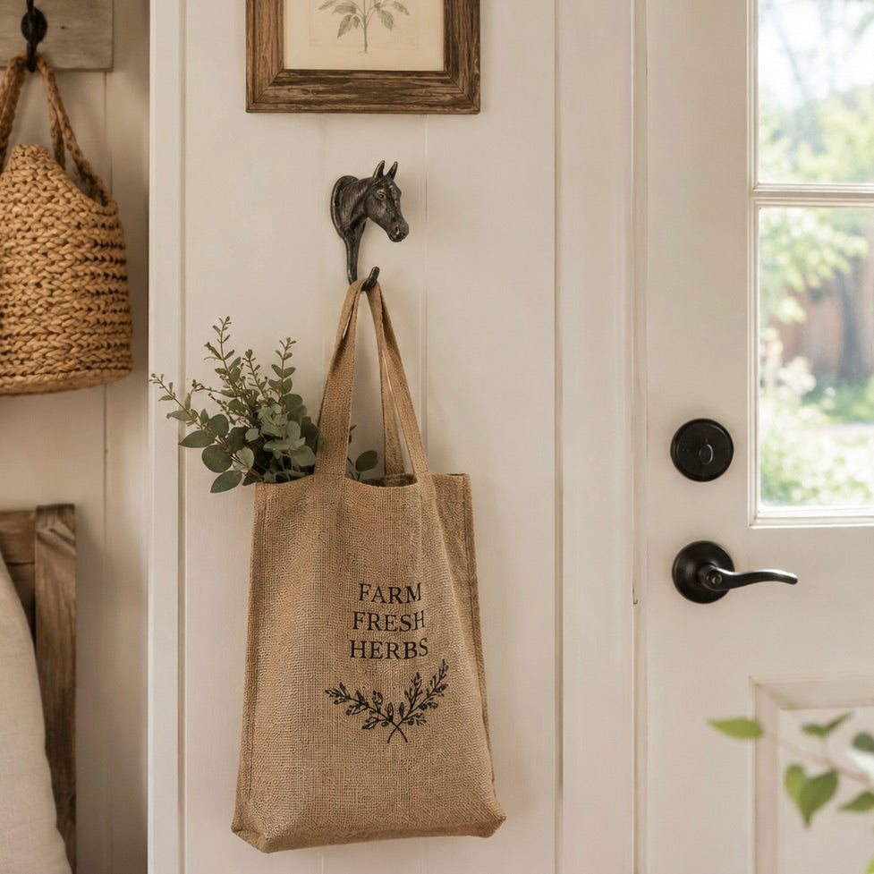 A natural jute tote bag with 'FARM FRESH HERBS' printed in black hangs from a horse-head door hook on a white paneled door. The bag contains greenery and is styled in a cozy, rustic entryway.