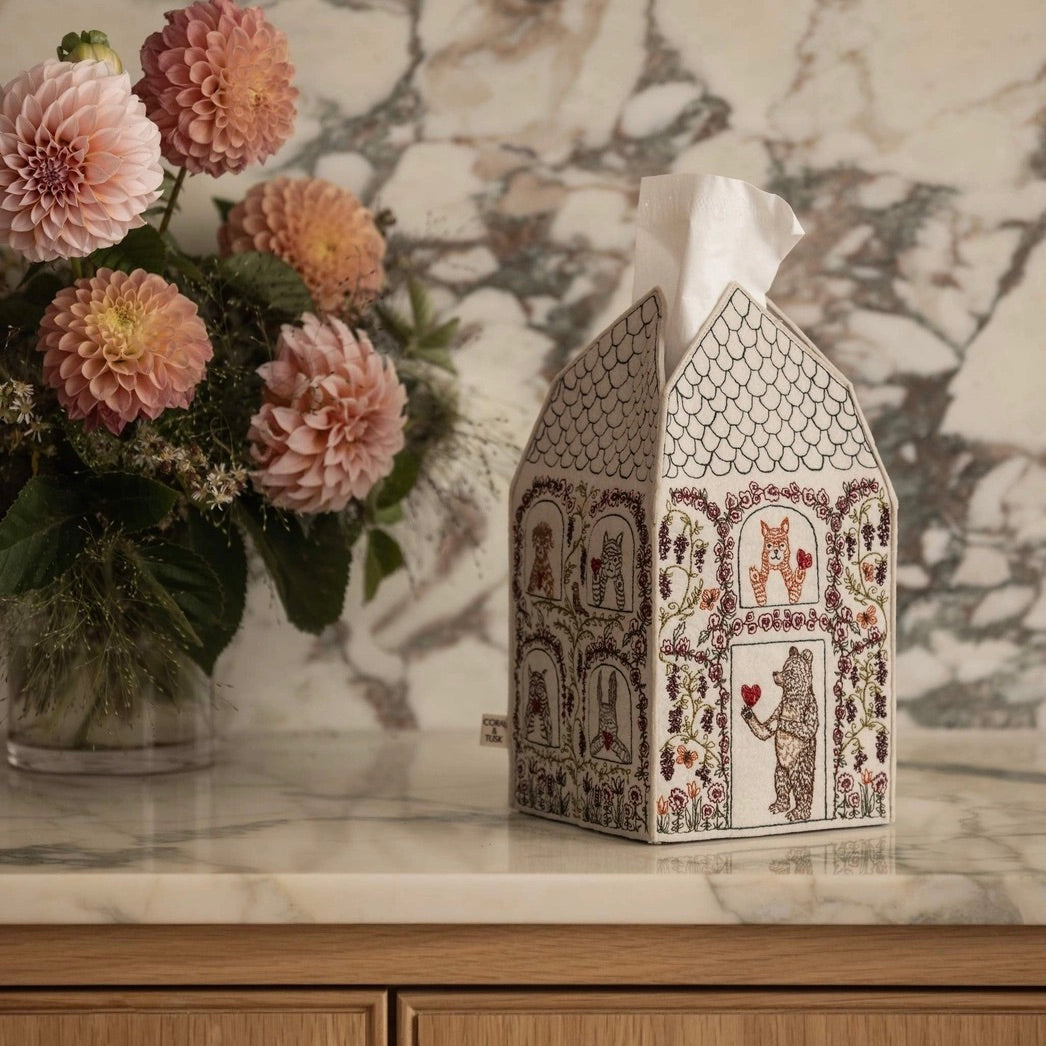 A decorative tissue box cover shaped like a house, printed with whimsical animal and floral illustrations, sits on a marble countertop beside a vase of pink flowers. A white tissue is partially visible at the top.
