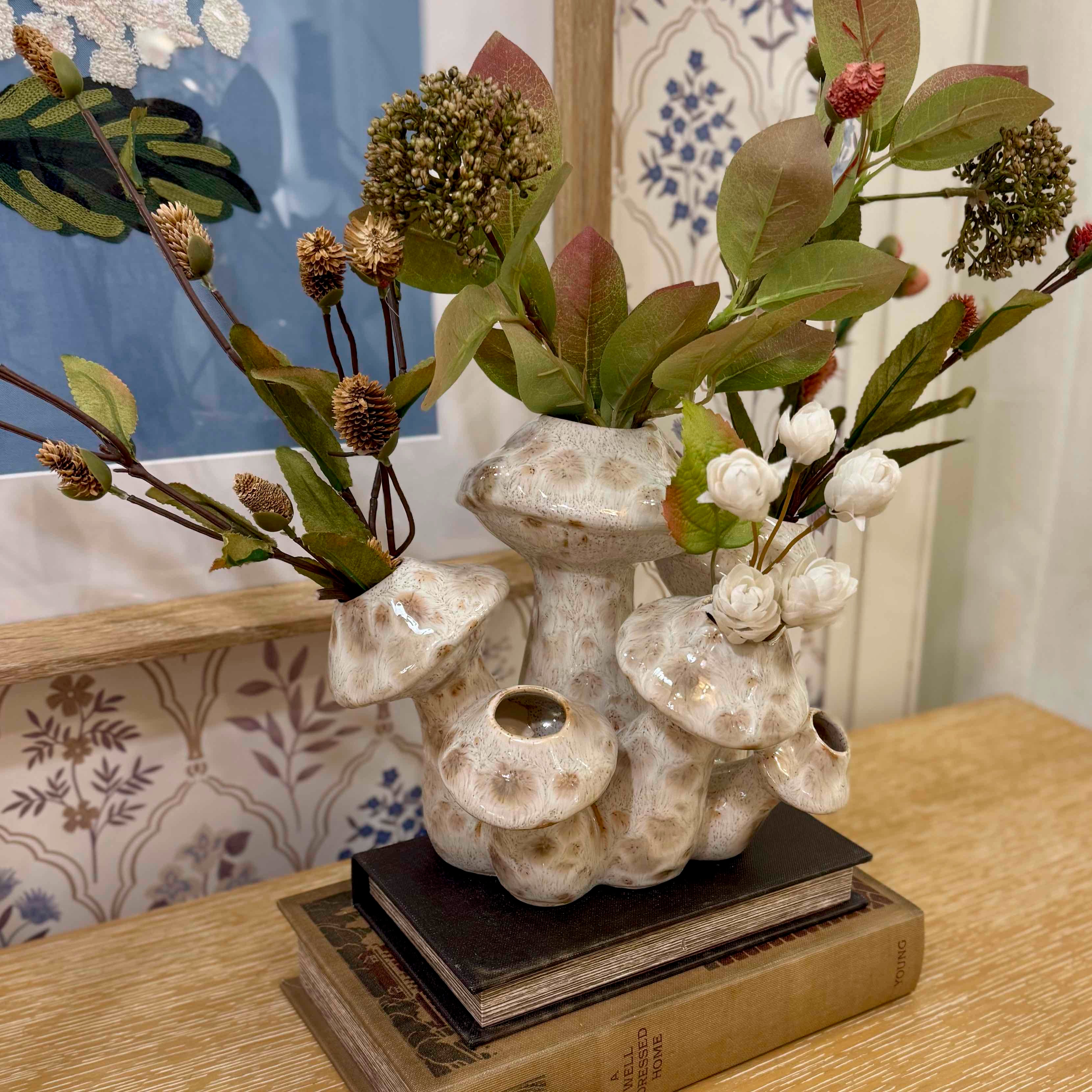 Decorative arrangement with plants and a textured vase on books against a patterned wall.
