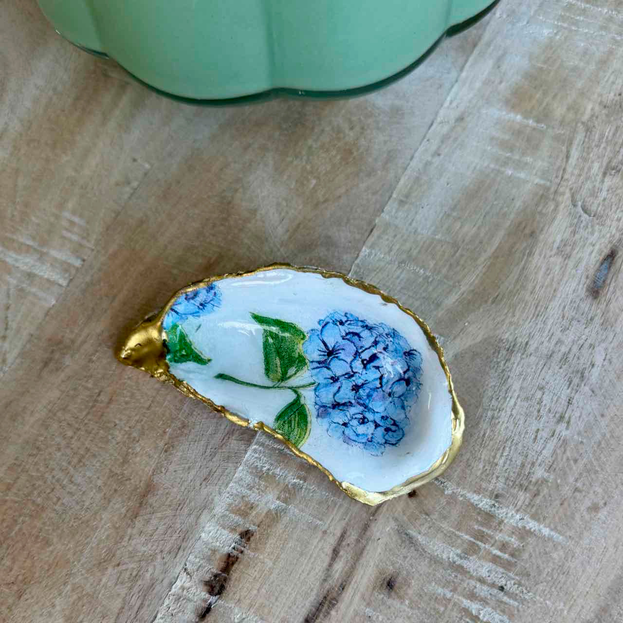 A small teardrop-shaped white dish with a gold-trimmed edge, decorated with blue flowers and green leaves, resting on a light wood surface.