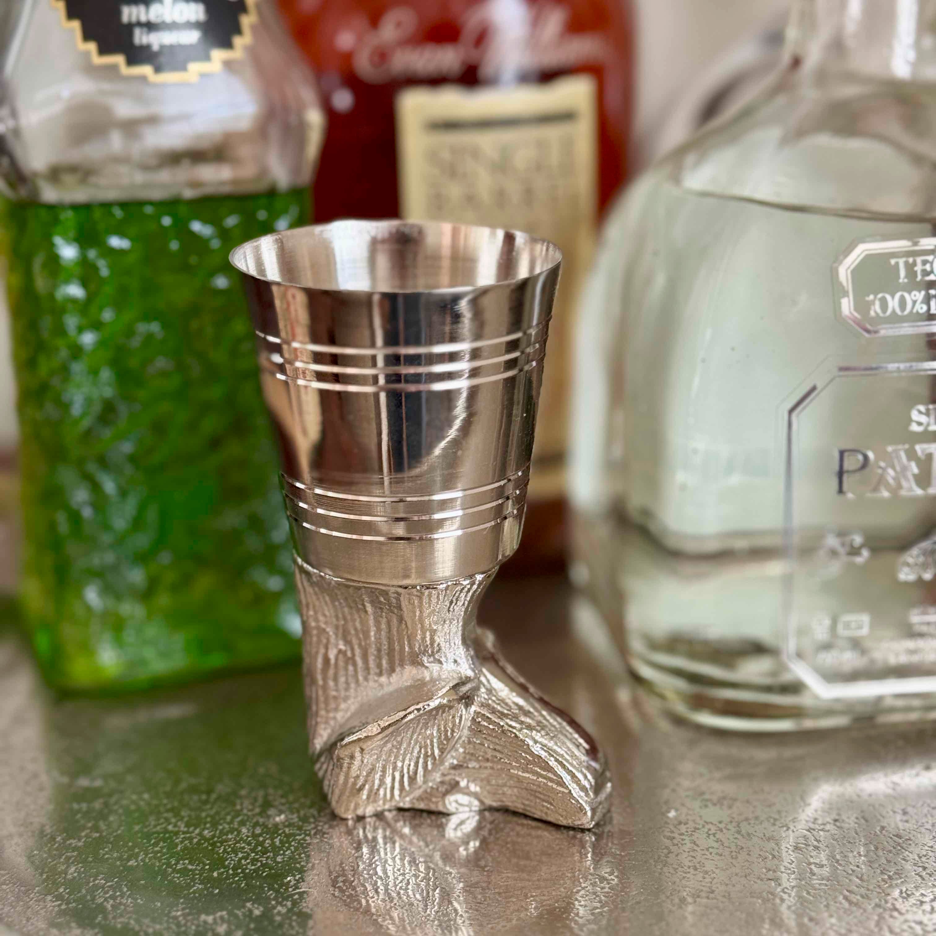 Silver boot-shaped jigger with bottles of tequila in the background