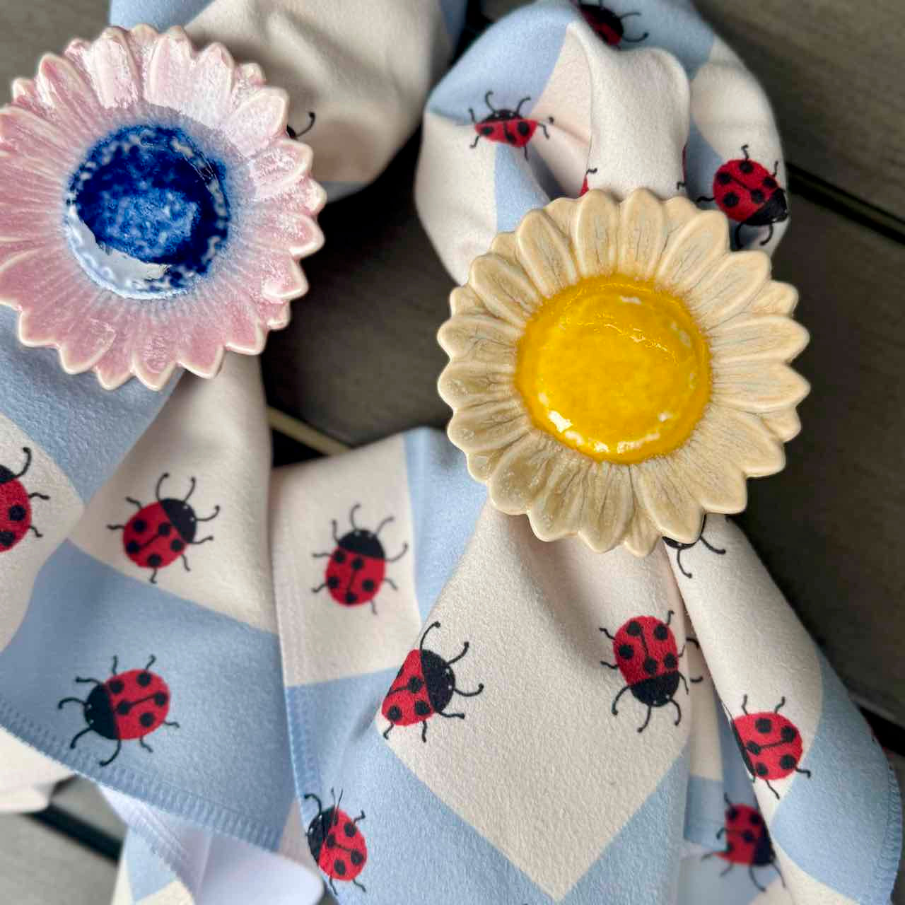 A close-up of a light blue and white checkered fabric napkin with a red ladybug print, tied with a bow and decorated with two ceramic daisy ring clips, one pink and one cream with a yellow center.