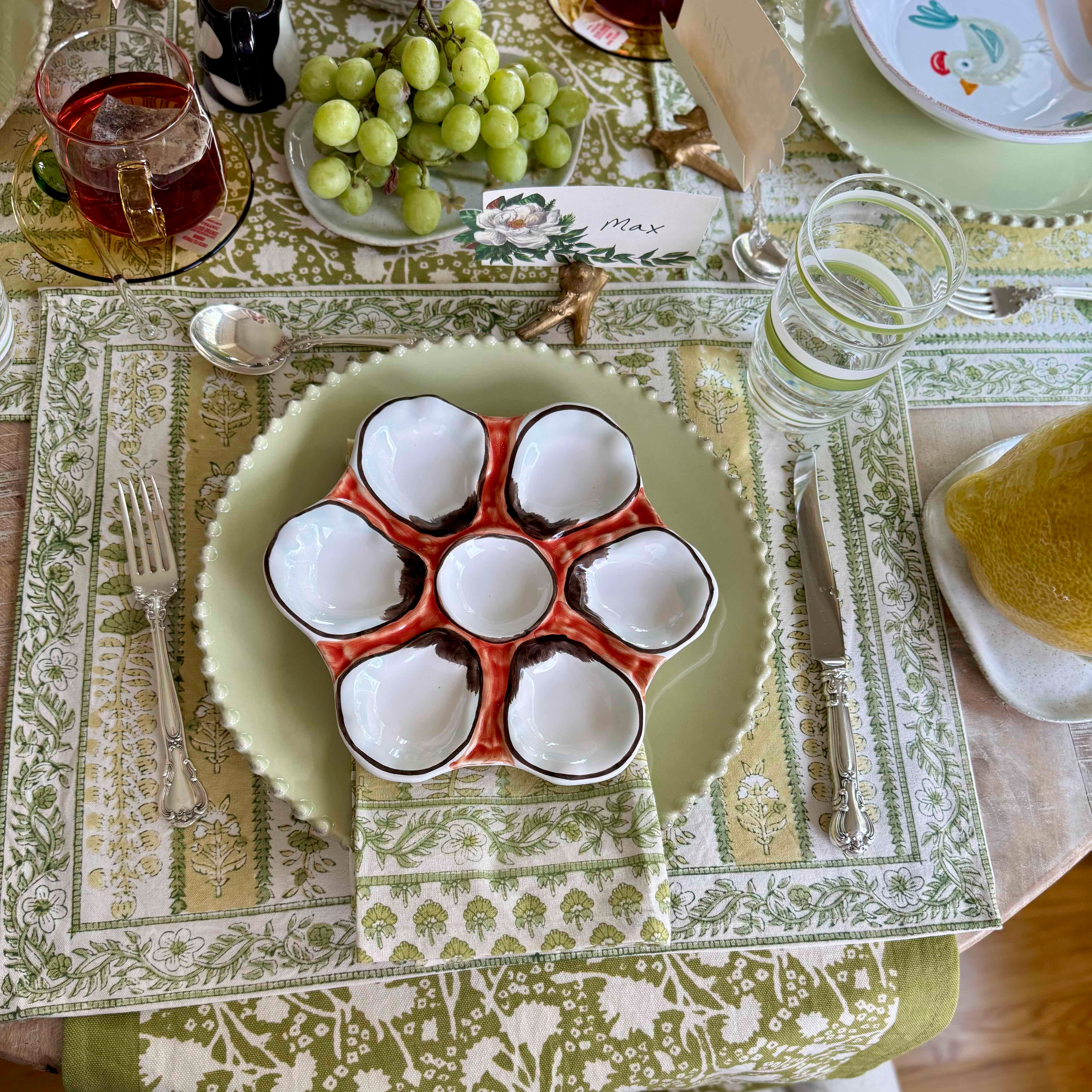 Eggs in a red holder on a green plate with a patterned tablecloth and glasses of juice in the background.