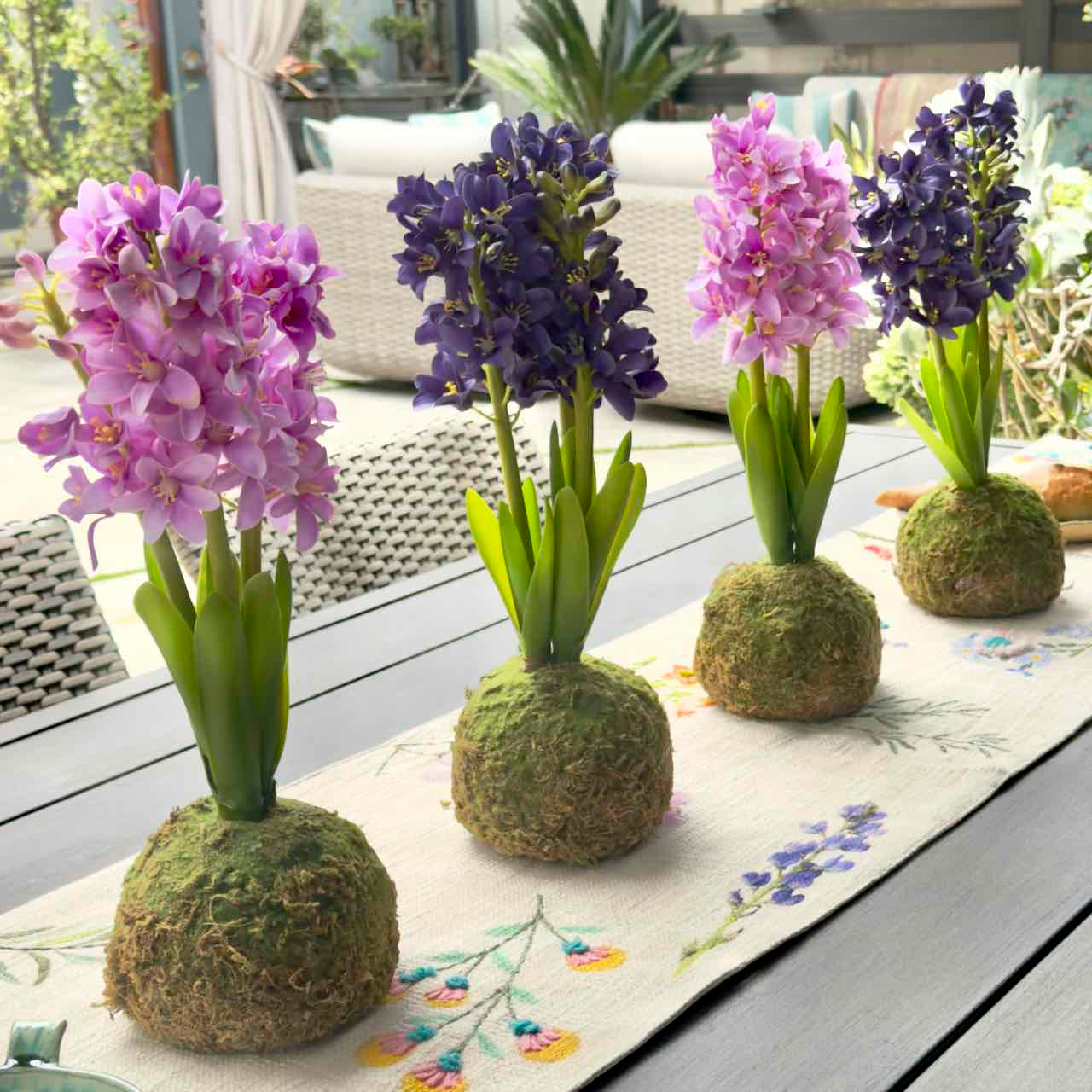 Four kokedama flower arrangements with purple and pink hyacinths in moss balls, displayed on a floral table runner on a gray wooden outdoor table.