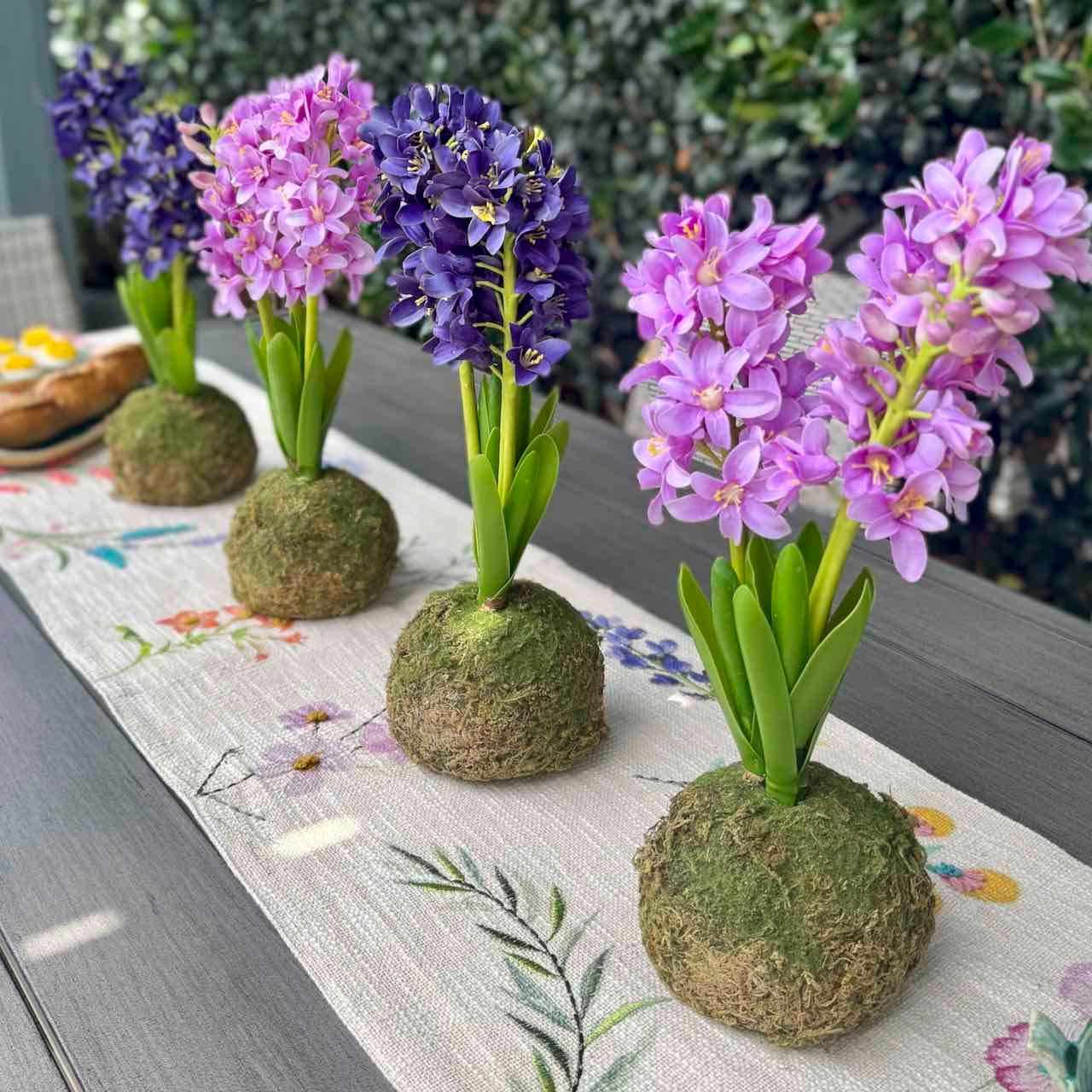 Five artificial hyacinth table centerpieces in purple and blue shades, each mounted in a mossy brown kokedaku ball on a white floral runner over a gray wooden table.