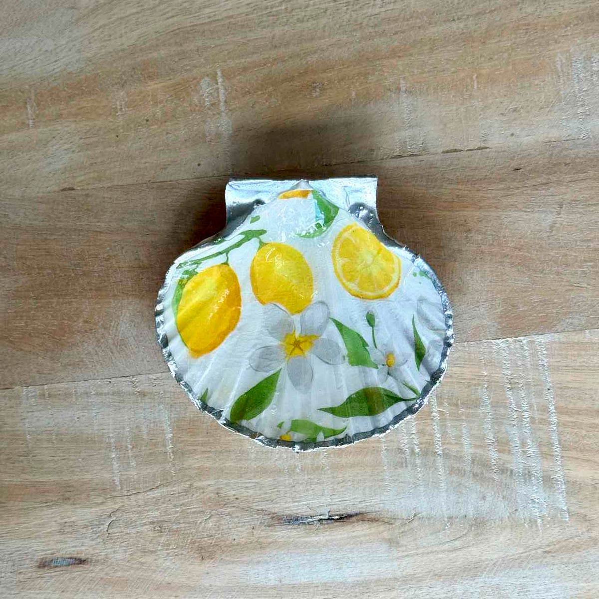 A scallop-shaped metal tin lid featuring a lemon and white flower pattern, resting on a light brown wooden surface.