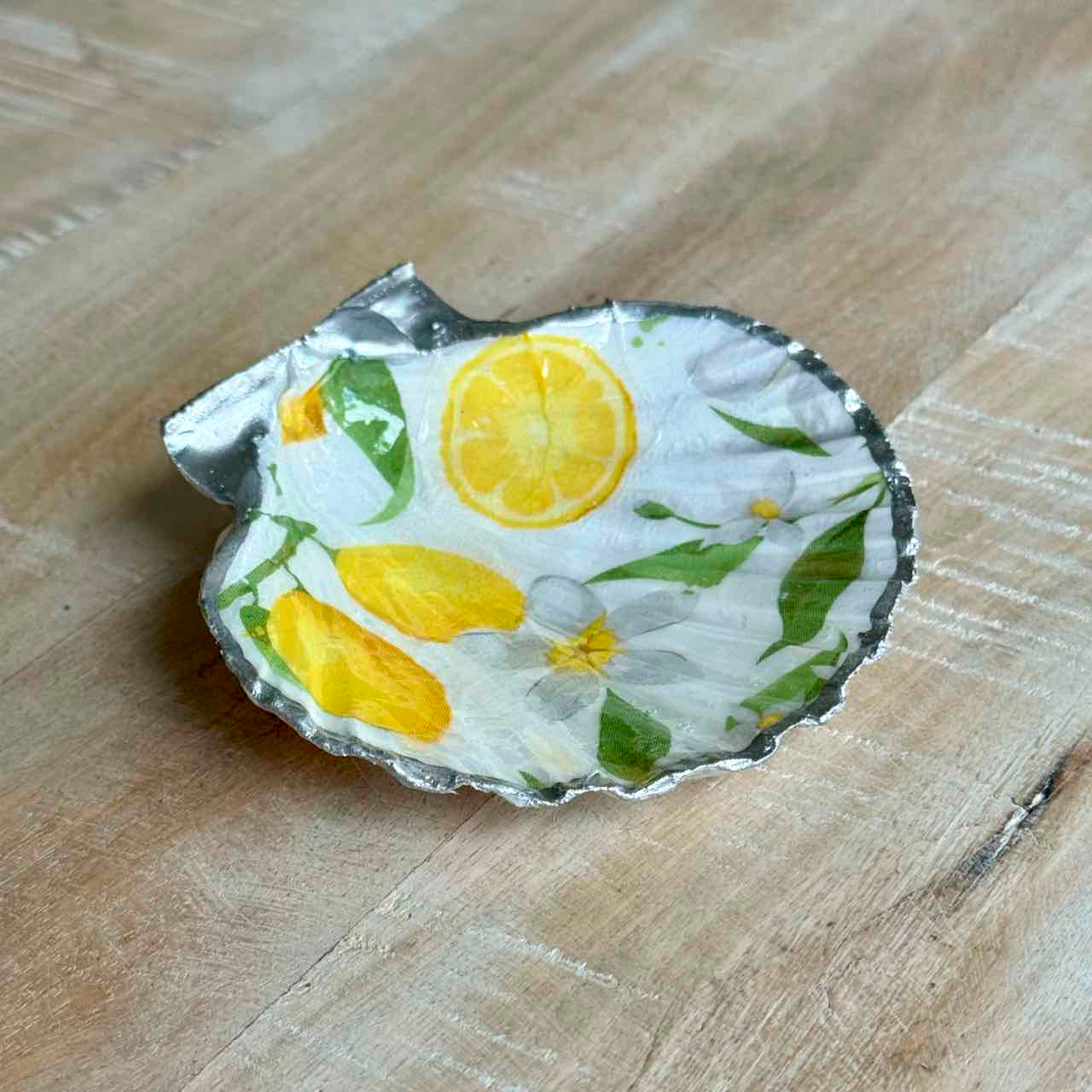 A small decorative dish made from a crushed aluminum can, decorated with a hand-painted lemon and flower pattern, resting on a light wood surface.
