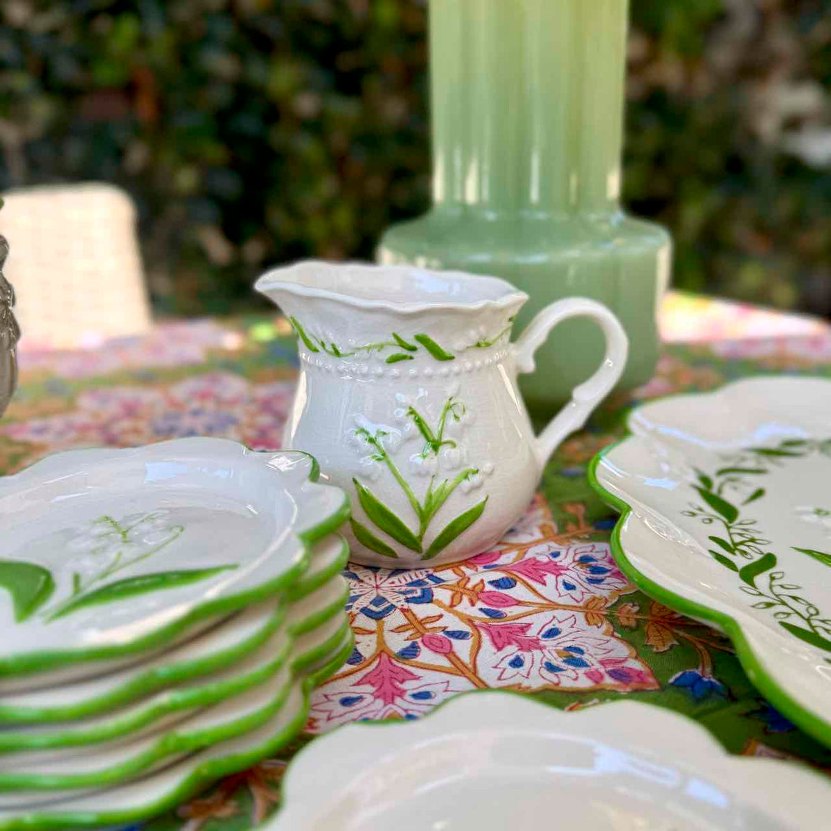 A white ceramic pitcher and matching plates with green leaf and floral patterns, arranged on a colorful floral tablecloth with a green glass vase in the background.