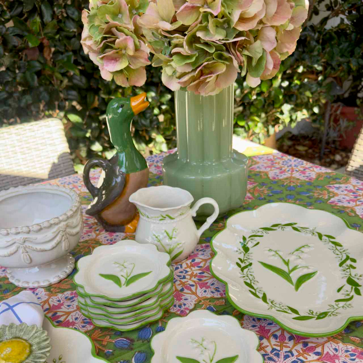 A styled collection of ceramic tableware displayed outdoors on a floral tablecloth, featuring a green duck-shaped pitcher, a ribbed light green vase with pink hydrangea flowers, a white embossed bowl, a cream pitcher with a green handle, and multiple white plates and bowls decorated with green floral and lily-of-the-valley motifs.