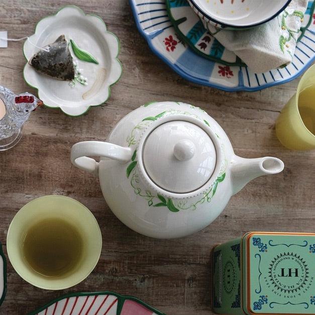 A white ceramic teapot with green floral vine patterns, styled on a rustic wooden table with matching tea cups, a teabag, and a decorative tea tin.