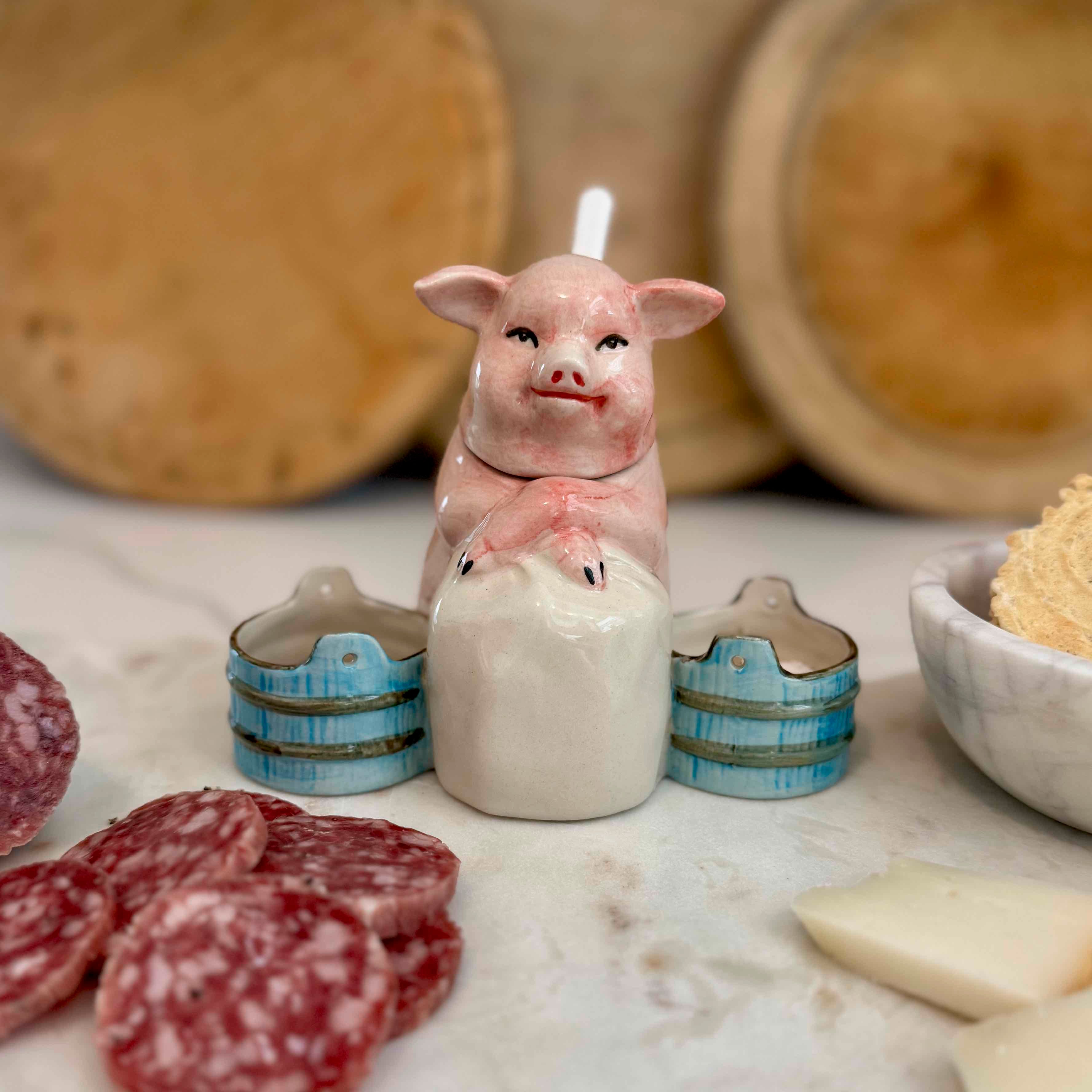 Ceramic pig candle with small barrels on a marble surface with cured meats and bread in the background.